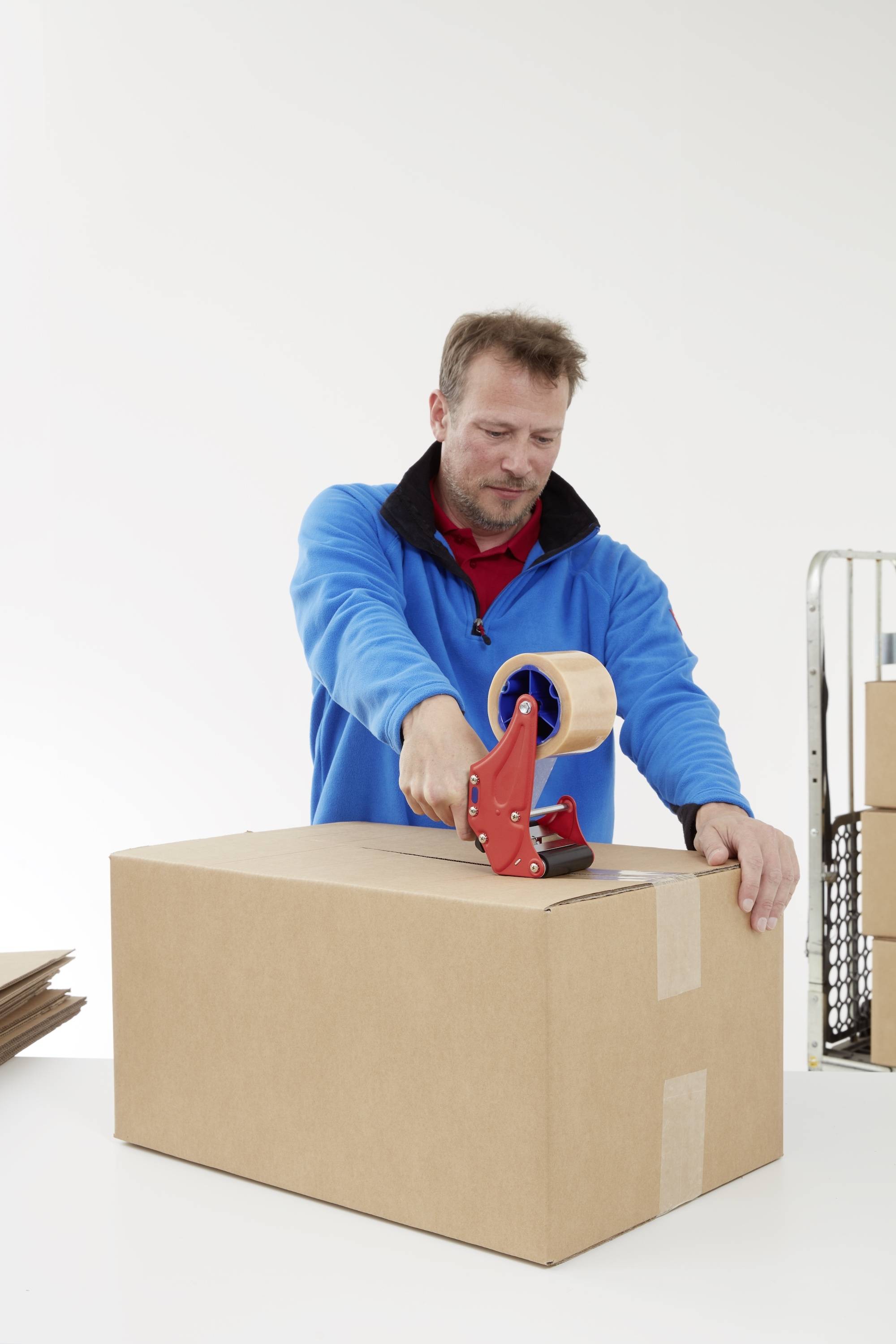 Ein Mann in blauer Jacke verschließt einen Karton mit Klebeband. Im Hintergrund sind weitere Kartons zu sehen.