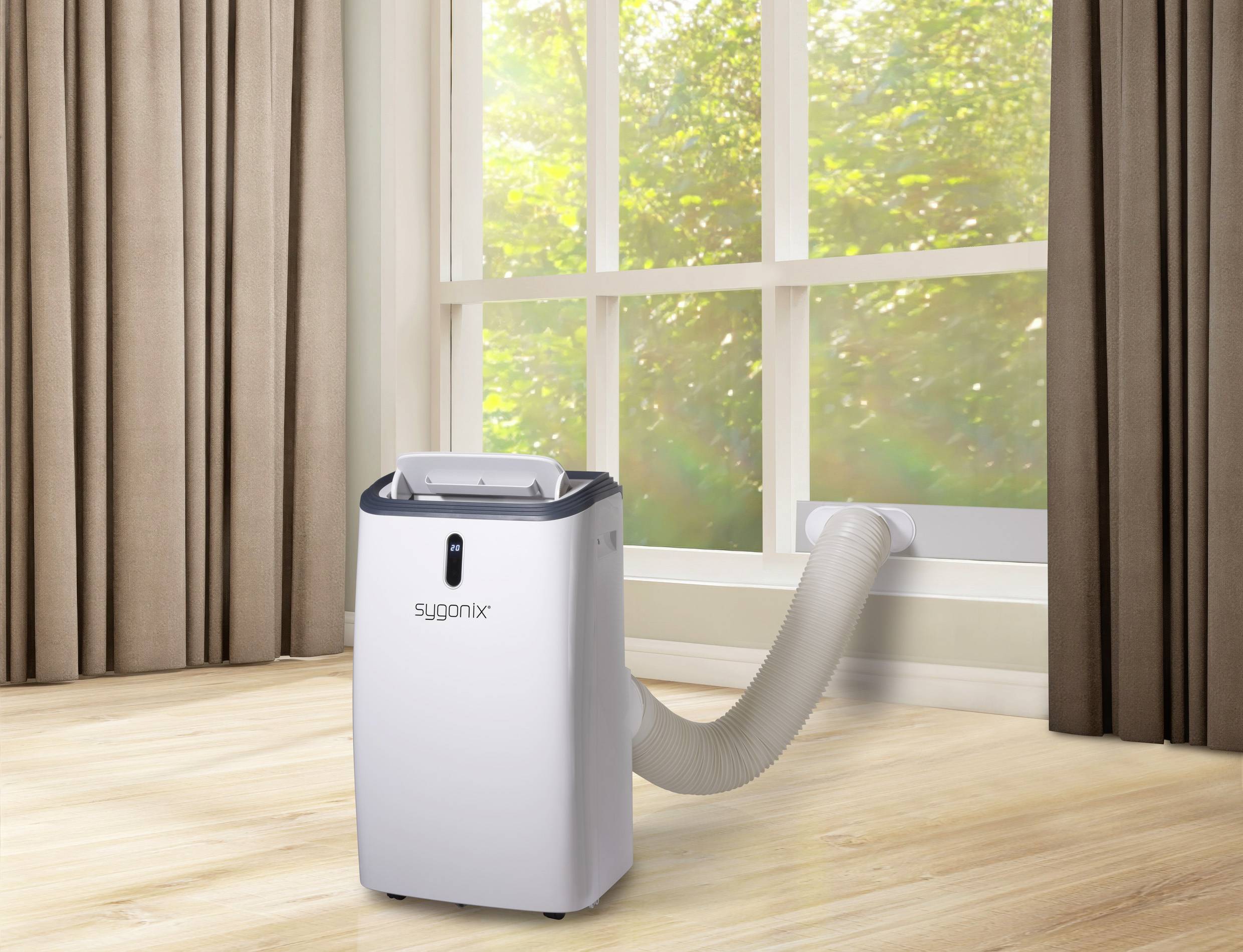 Ein tragbares Klimagerät steht in einem Raum mit braunen Vorhängen und einem Fenster. Ein Schlauch führt aus dem Gerät nach draußen.