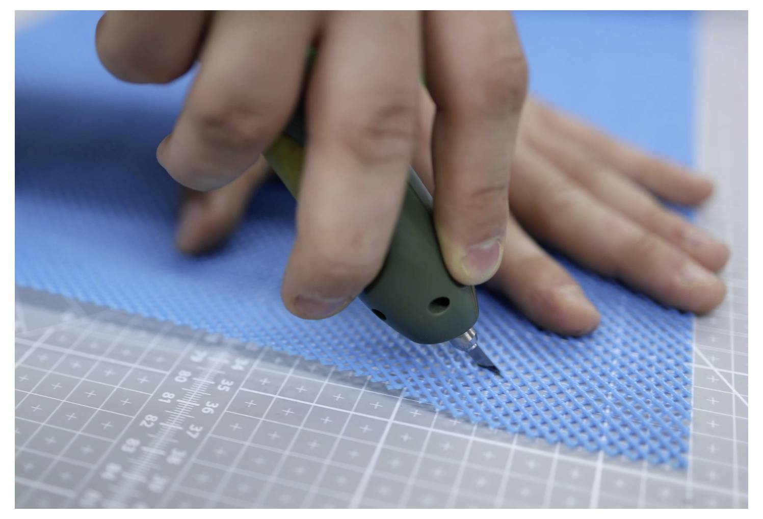 Eine Person schneidet ein Stück blaues strukturiertes Material mit einem Universalmesser auf einer Rastertechniker-Unterlage und zeigt dabei präzise Handwerksarbeit.