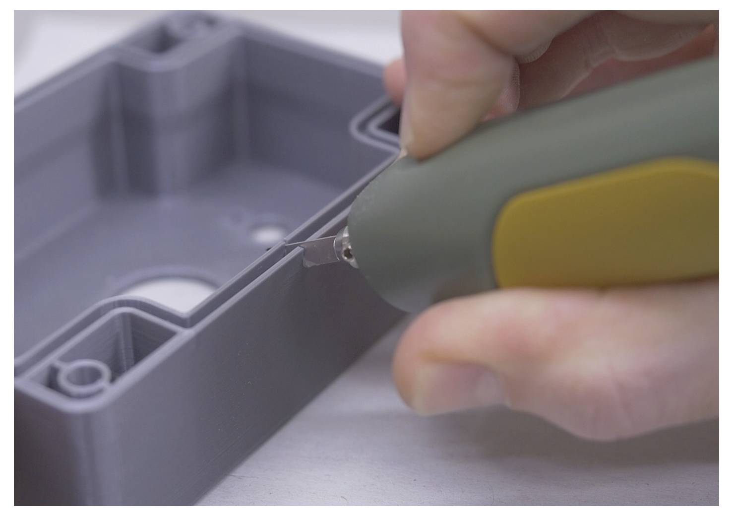 Eine Hand, die ein grün-gelbes Utility-Messer verwendet, um präzise und detailliert in den Rand eines grauen Kunststoffgehäuses zu schneiden.