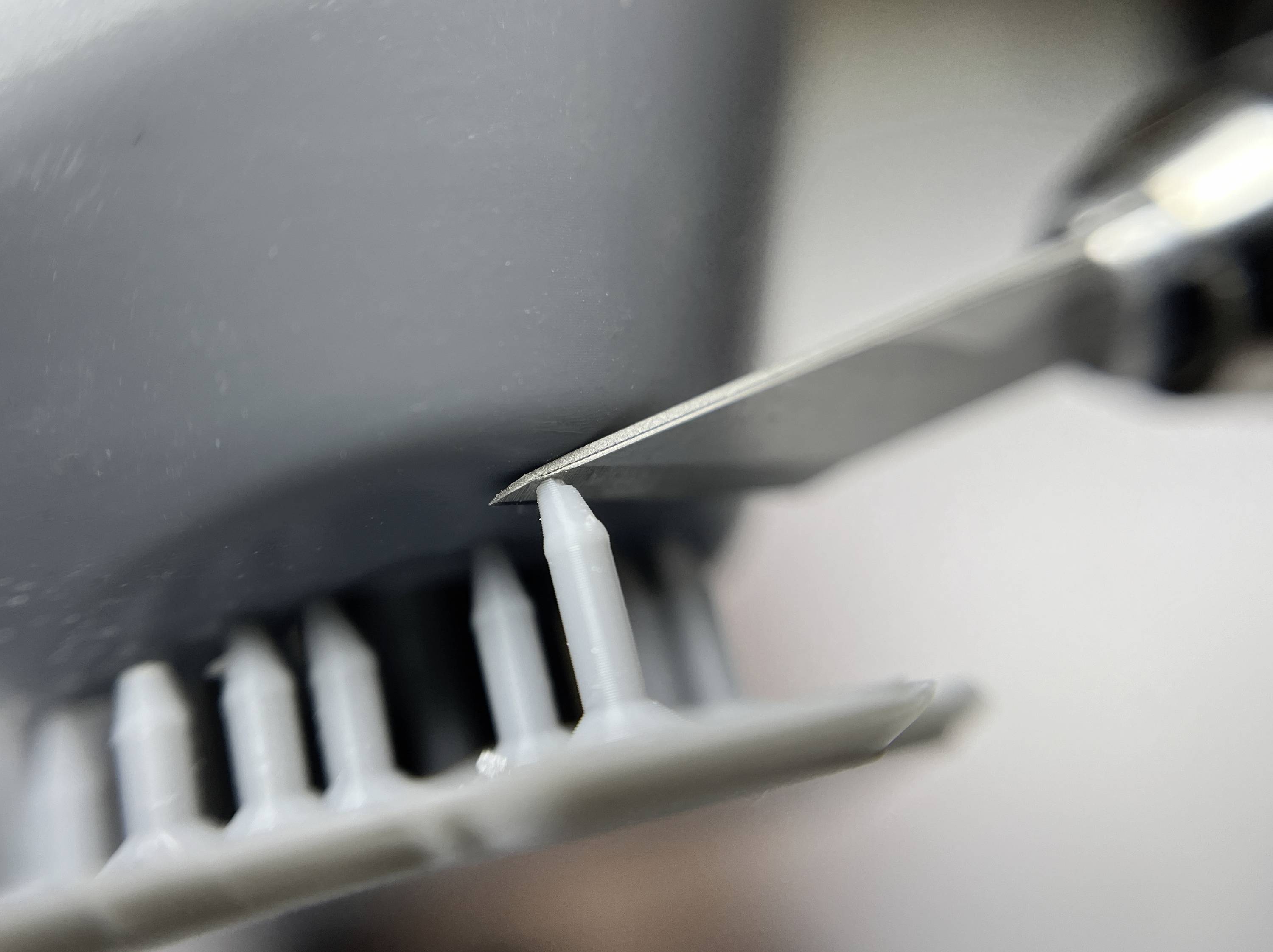 Eine Nahaufnahme einer scharfen Klinge, die überschüssiges Plastik von einem grauen Modellteil sorgfältig abschneidet und präzise Arbeit beim Modellbau zeigt.