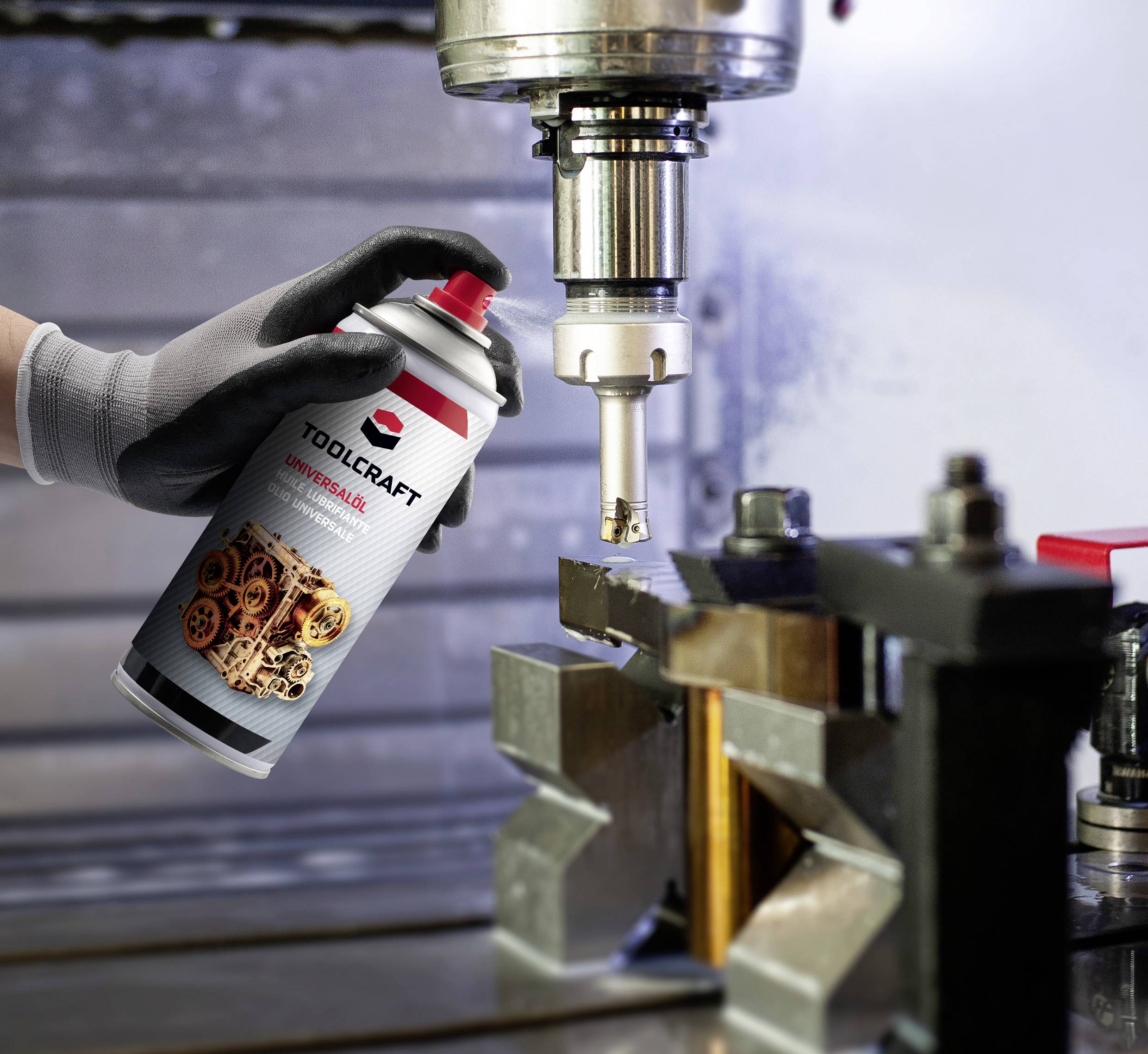 Eine Hand hält eine Spraydose mit Schmiermittel, richtet sie auf eine metallene Fräsmaschine. Im Hintergrund sind Werkzeuge und Maschinen erkennbar.