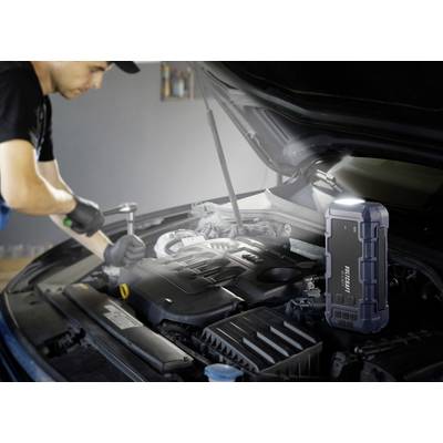 Ein Mechaniker arbeitet am Motor eines Autos mit geöffnetem Motorraum. Im Vordergrund steht ein tragbares Starthilfegerät.