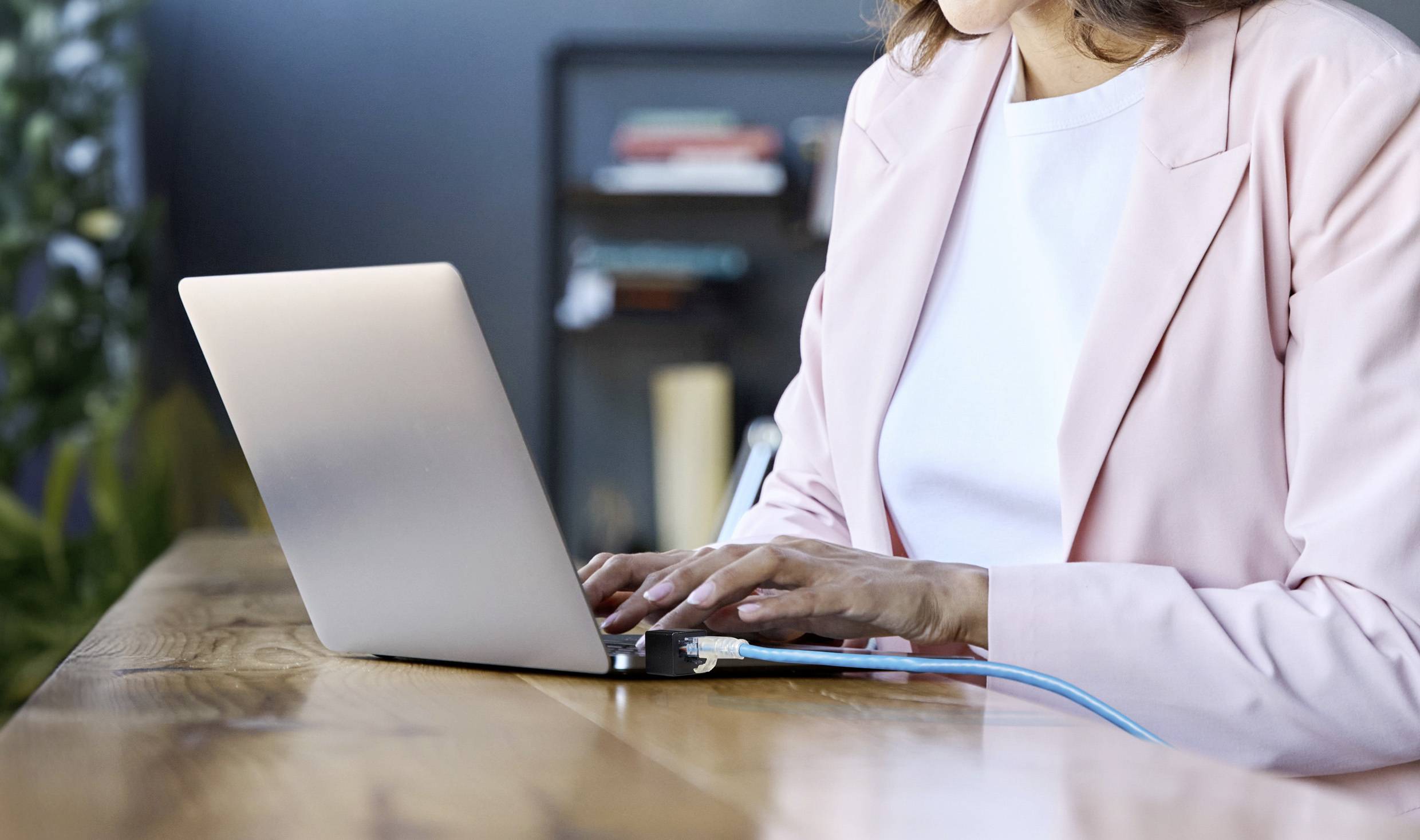 Eine Person in einem rosa Blazer arbeitet an einem Laptop auf einem Holztisch. Im Hintergrund sind Bücher und Pflanzen sichtbar.
