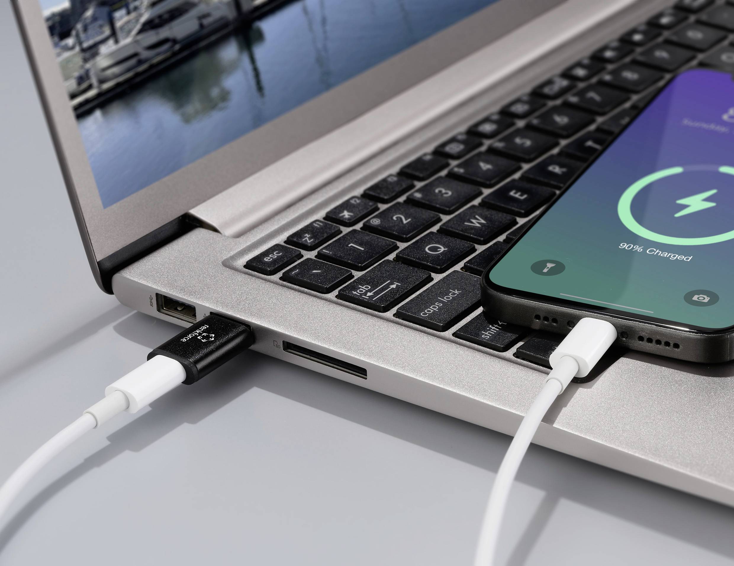 Ein Smartphone wird über ein Kabel an einen Laptop angeschlossen und zeigt '90% geladen' an. Im Hintergrund sind Boote zu sehen.