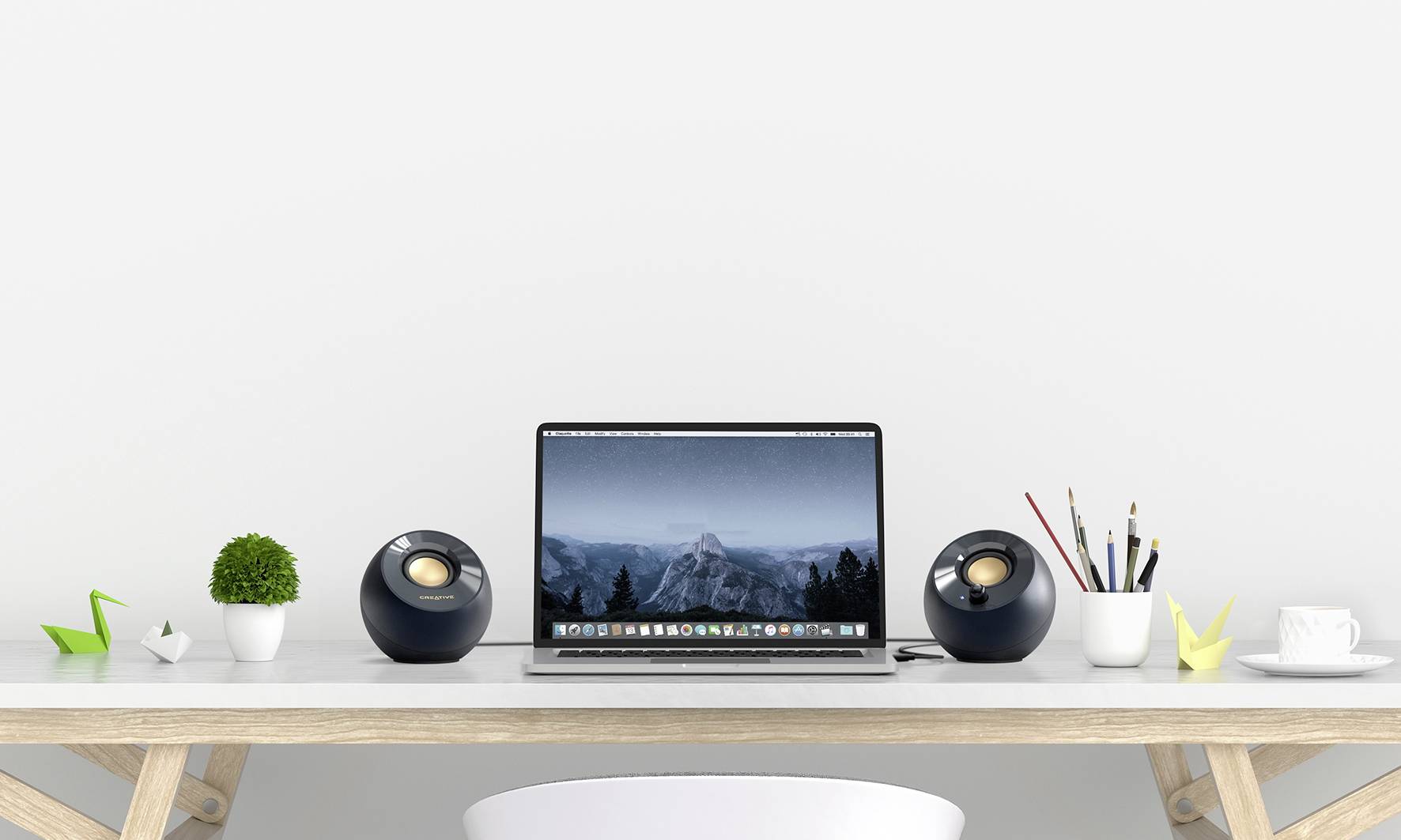 Ein ordentlicher Schreibtisch mit einem Laptop in der Mitte, umgeben von Lautsprechern, Pflanze, Stiften und Papierkram.