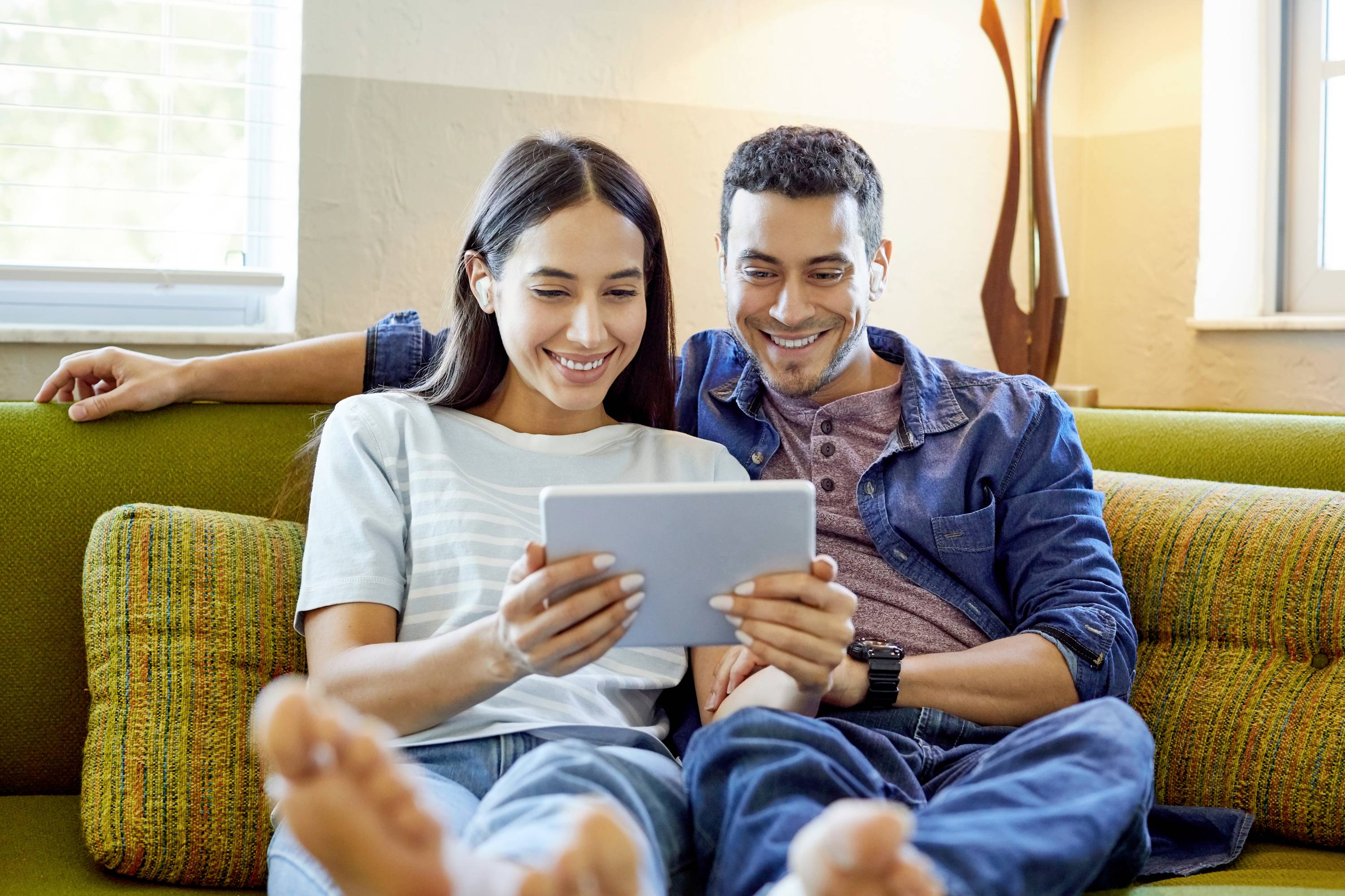 Ein Paar sitzt entspannt auf einem Sofa und lächelt, während es ein Tablet gemeinsam ansieht. Hintergrund zeigt eine Lampe und Fenster.
