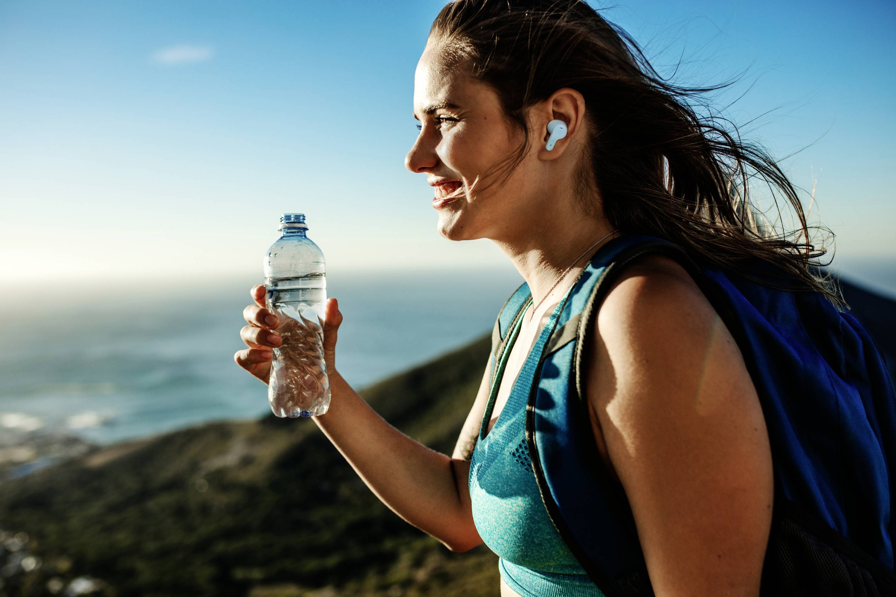 Frau beim Wandern auf einem Bergpfad mit Wasserflasche in der Hand; sonniger Tag, Natur im Hintergrund.