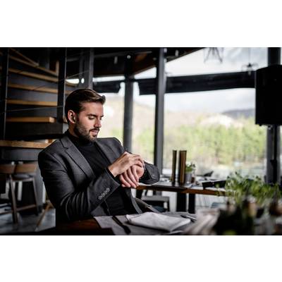 Ein Mann in einem Anzug sitzt in einem modernen Restaurant am Tisch und schaut auf seine Armbanduhr. Im Hintergrund ist Natur zu sehen.
