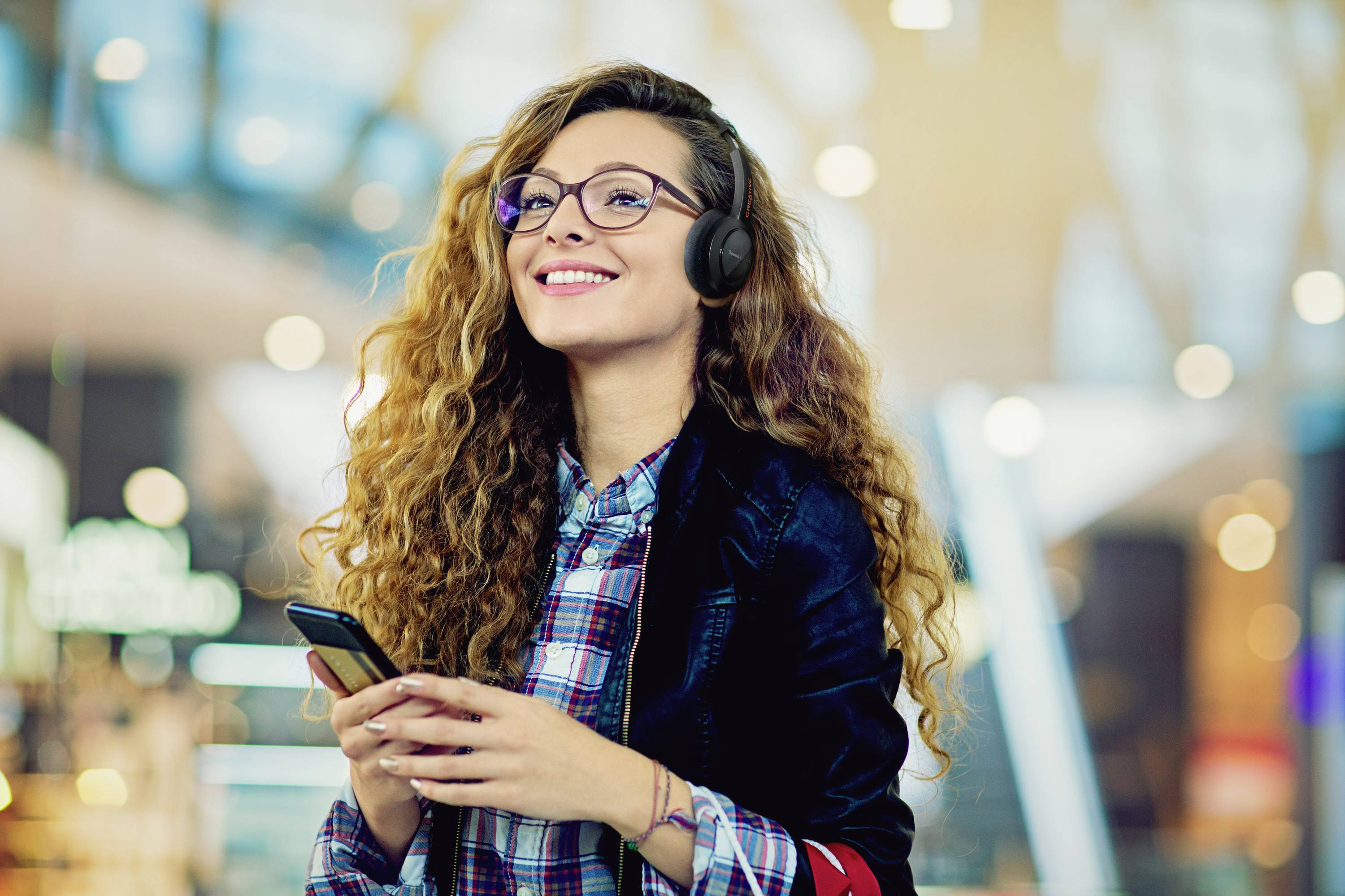 Junge Frau mit Brille und Kopfhörern lächelt, während sie ein Smartphone in der Hand hält. Hintergrund unscharf, belebt.