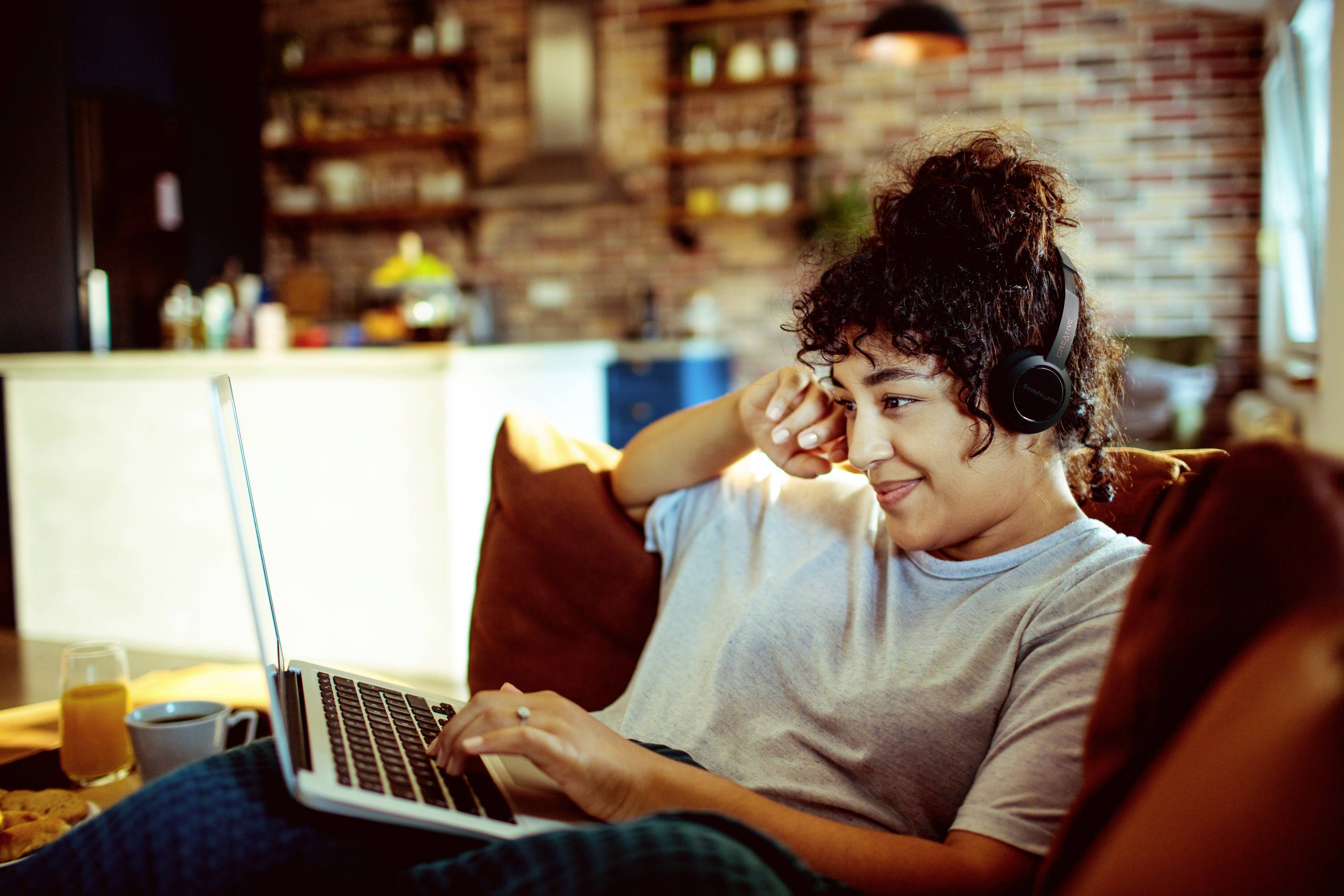 Eine Frau sitzt lächelnd auf einem Sofa, trägt Kopfhörer und tippt auf einem Laptop. Im Hintergrund ist eine Küche zu sehen.
