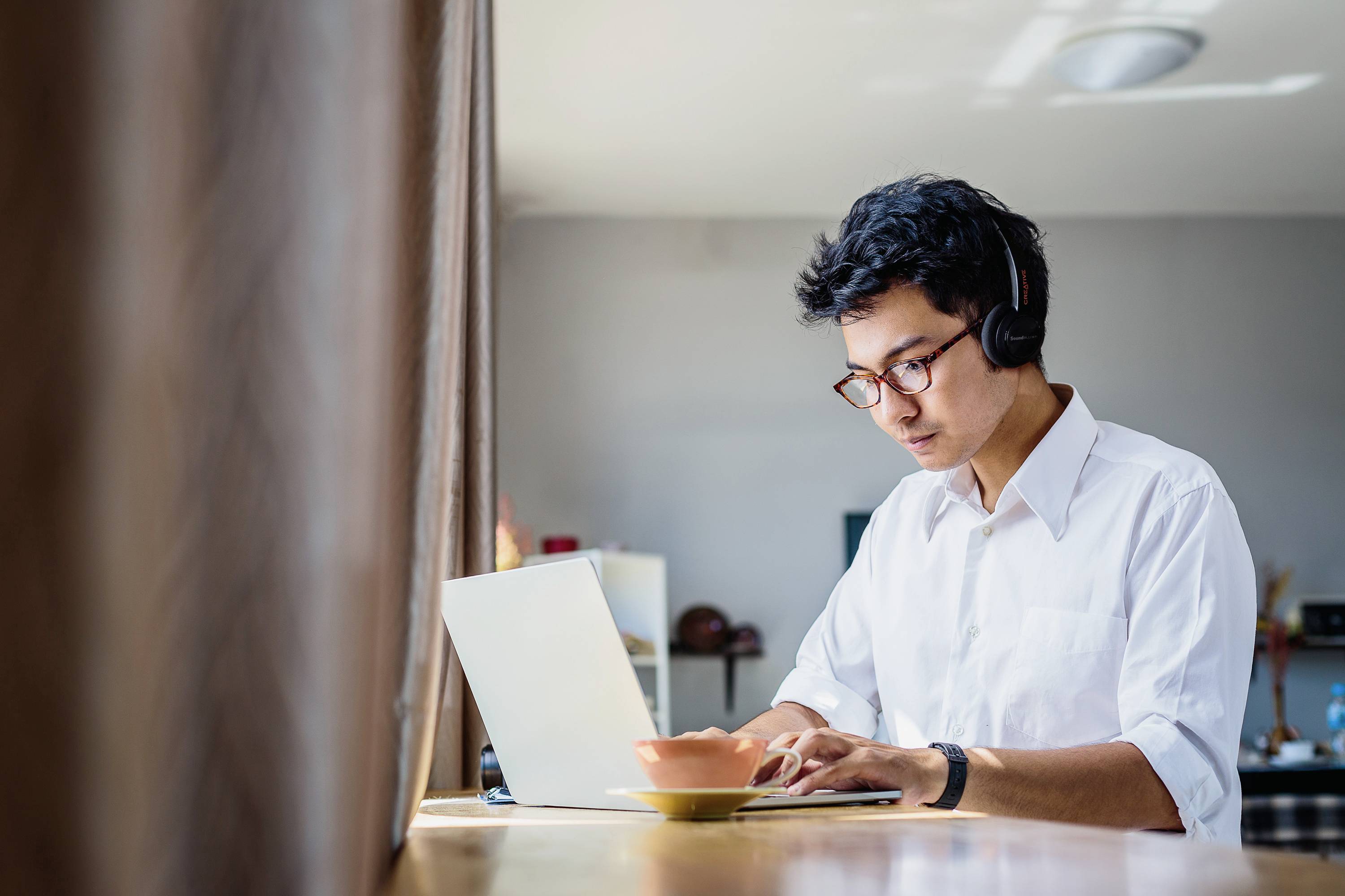 Ein Mann mit Brille arbeitet konzentriert an einem Laptop, trägt Kopfhörer und sitzt in einem hellen Raum. Auf dem Tisch steht eine Tasse.