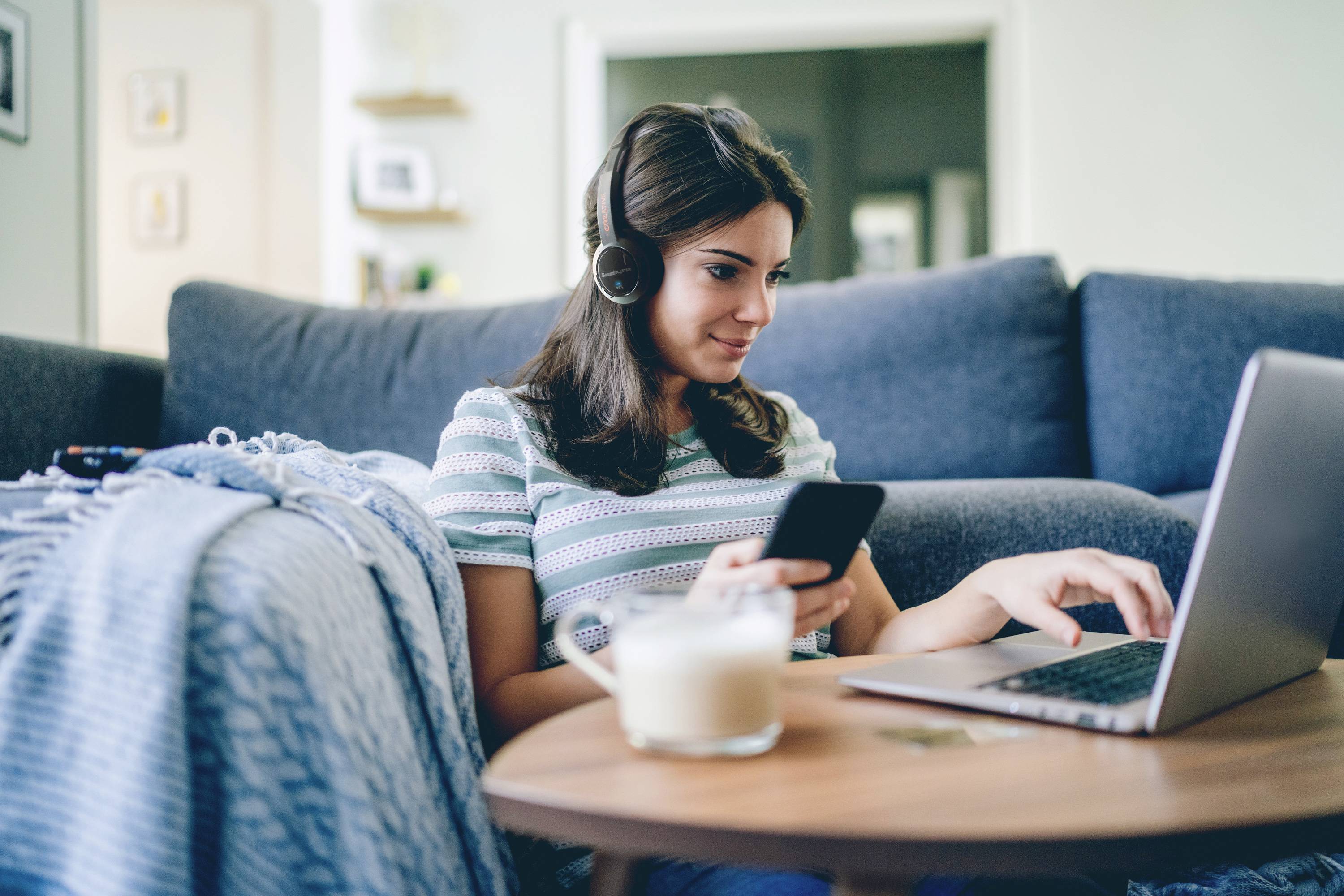 Eine Frau sitzt auf dem Sofa, trägt Kopfhörer und arbeitet am Laptop, während sie ein Smartphone hält. Im Vordergrund steht ein Tisch mit einem Getränk.