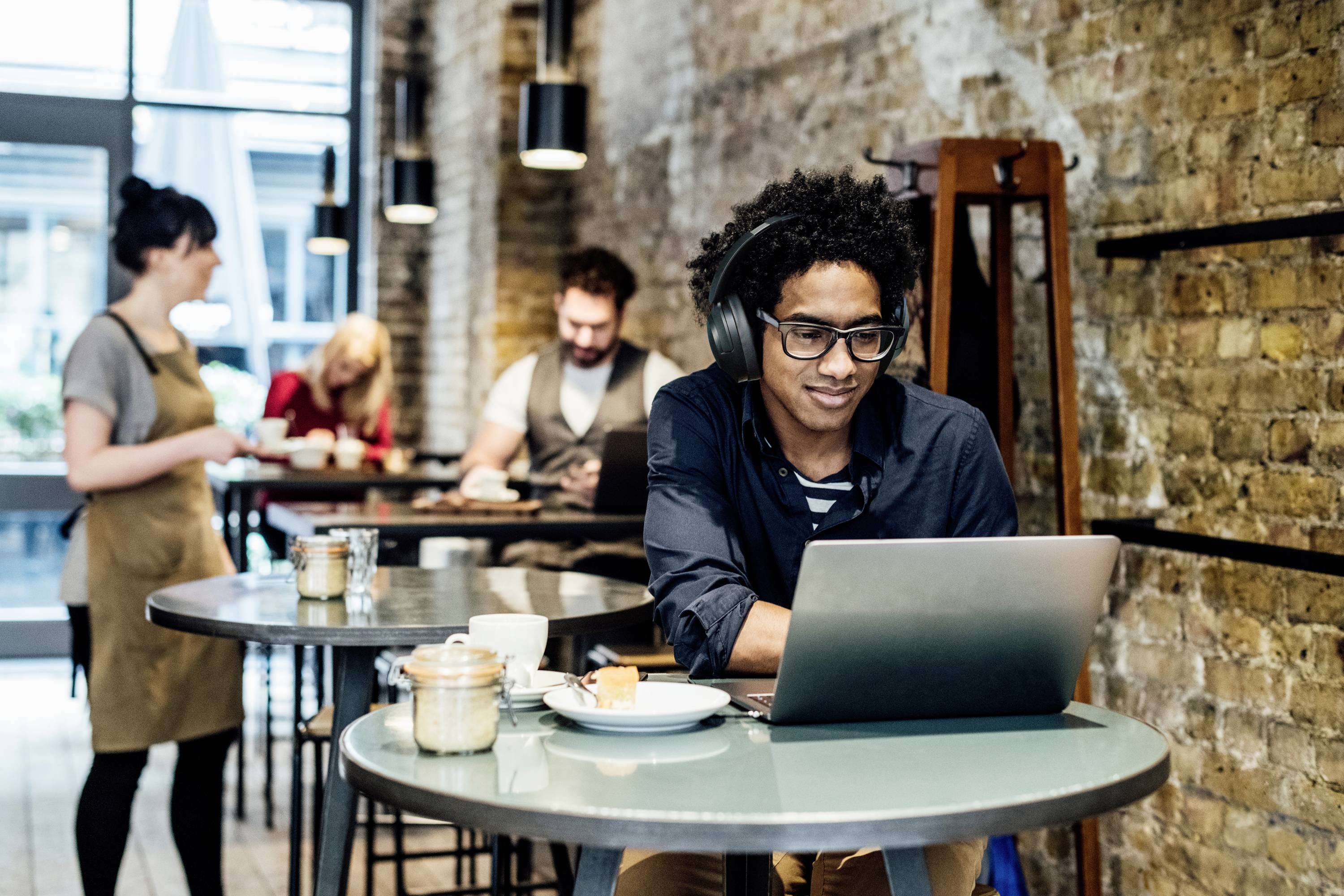 Ein Mann mit Kopfhörern arbeitet an einem Laptop in einem Café. Im Hintergrund bedienen zwei Mitarbeiter einen weiteren Kunden.