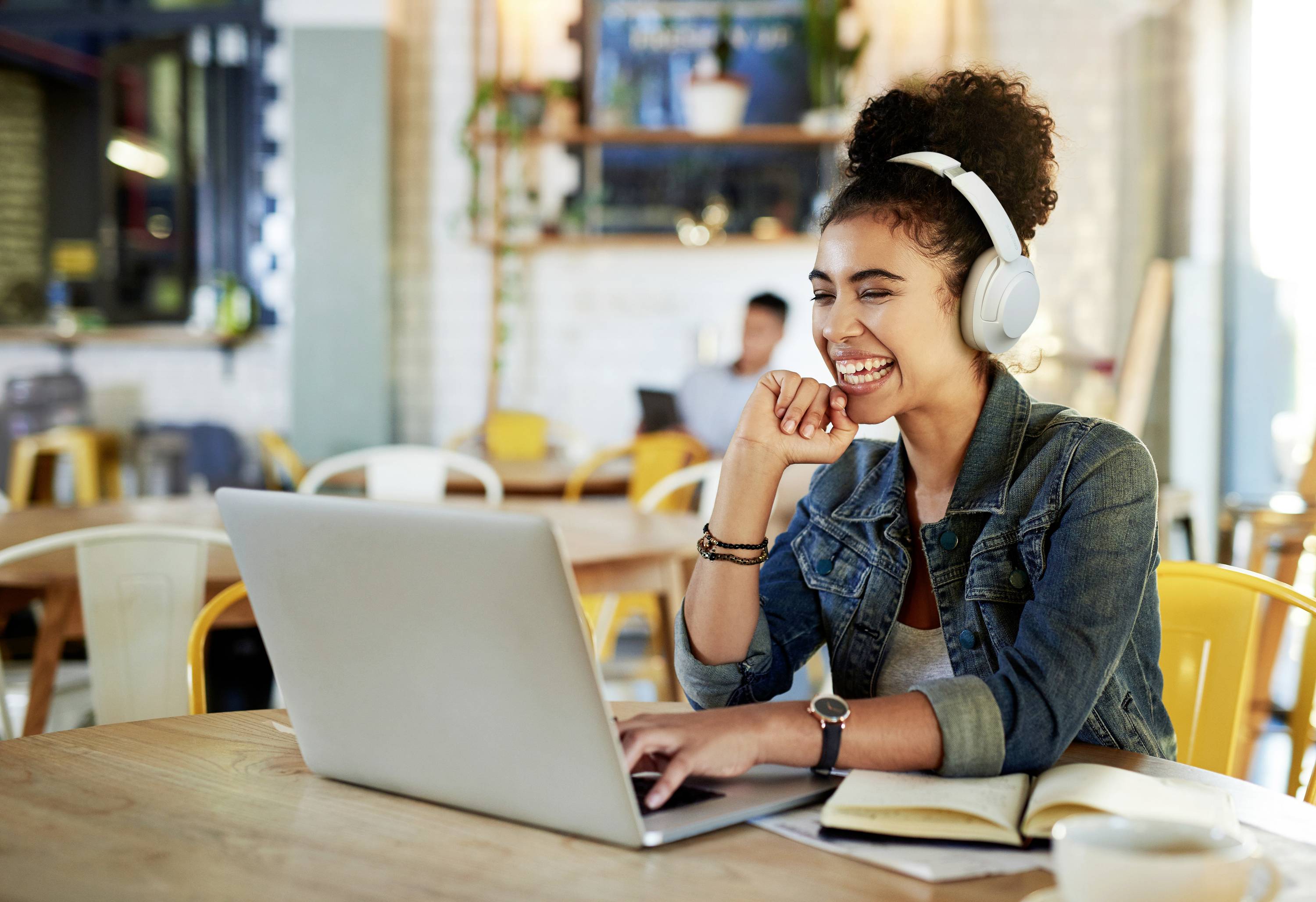 Eine lächelnde Person mit Kopfhörern sitzt in einem Café und arbeitet entspannt an einem Laptop.