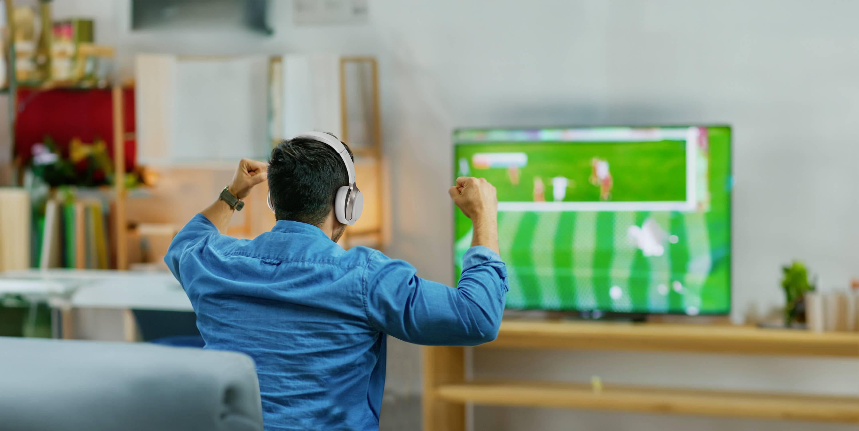 Ein Mann mit Kopfhörern sitzt auf einem Sofa und jubelt, während er ein Fußballspiel auf einem Fernseher in einem Wohnzimmer schaut.