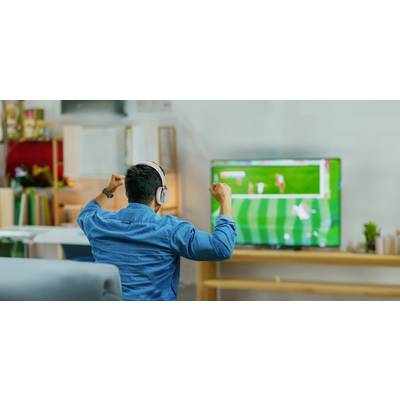 Ein Mann mit Kopfhörern sitzt auf einem Sofa und jubelt, während er ein Fußballspiel auf einem Fernseher in einem Wohnzimmer schaut.