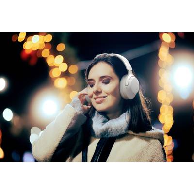 Eine lächelnde Person trägt Kopfhörer und genießt Musik, während sie nachts auf einem festlich beleuchteten Weihnachtsmarkt steht.