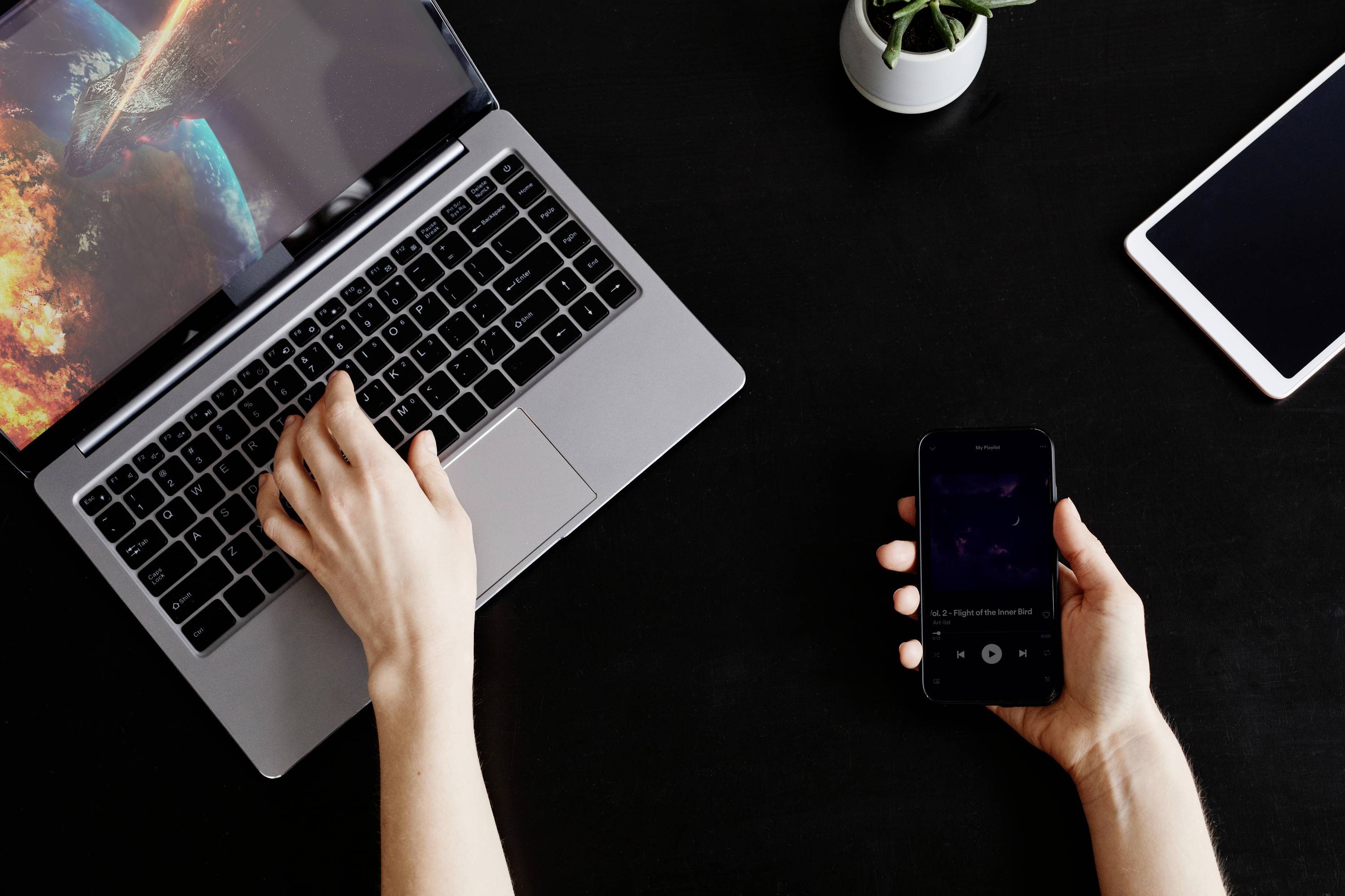 Eine Person tippt auf einem Laptop, während sie ein Smartphone in der anderen Hand hält. Ein Tablet liegt daneben auf dem Tisch.