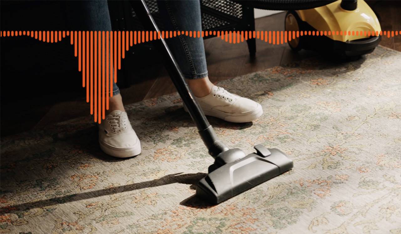 Person saugt einen Teppich mit einem Staubsauger. Ein gelbes Staubsaugergerät ist im Hintergrund. Eine Audiowellenform schwebt darüber.