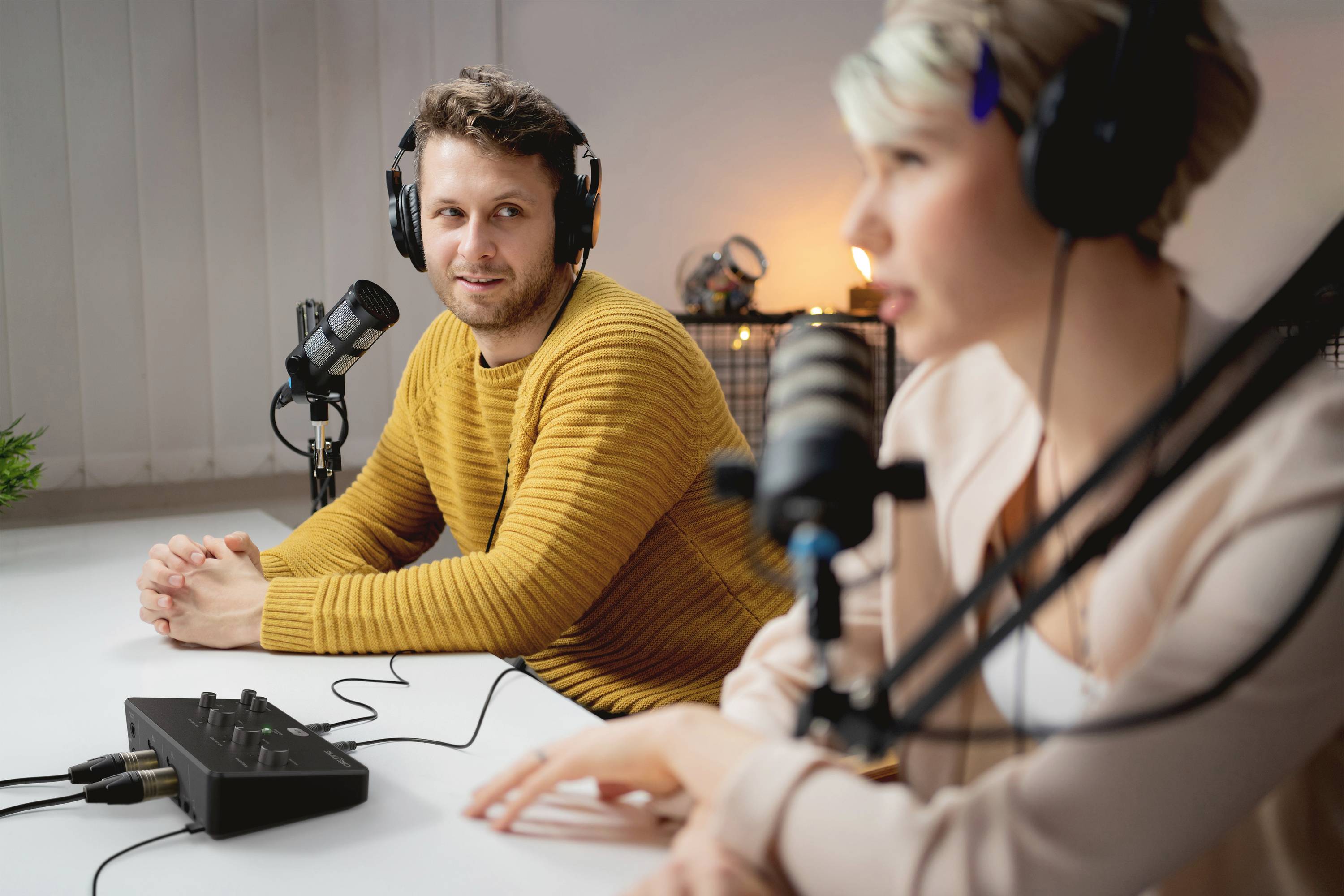 Zwei Personen nehmen an einem Tisch mit Mikrofonen und Kopfhörern eine Podcast- oder Radioaufnahme auf. Eine Person spricht, die andere hört zu.