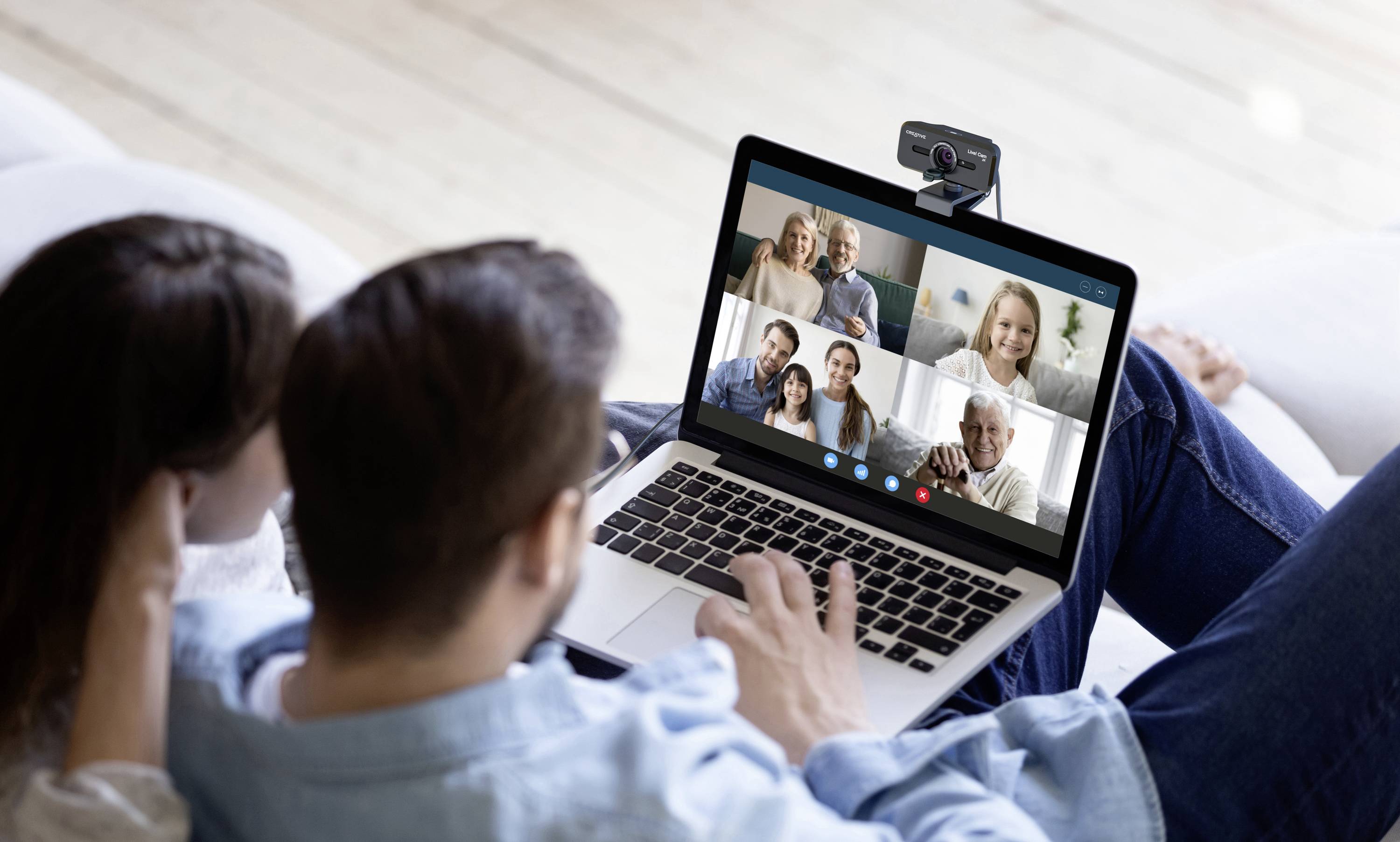 Eine Person auf einem Sofa nutzt einen Laptop für einen Videoanruf mit mehreren Teilnehmern. Die Webcam ist oben am Laptop angebracht.