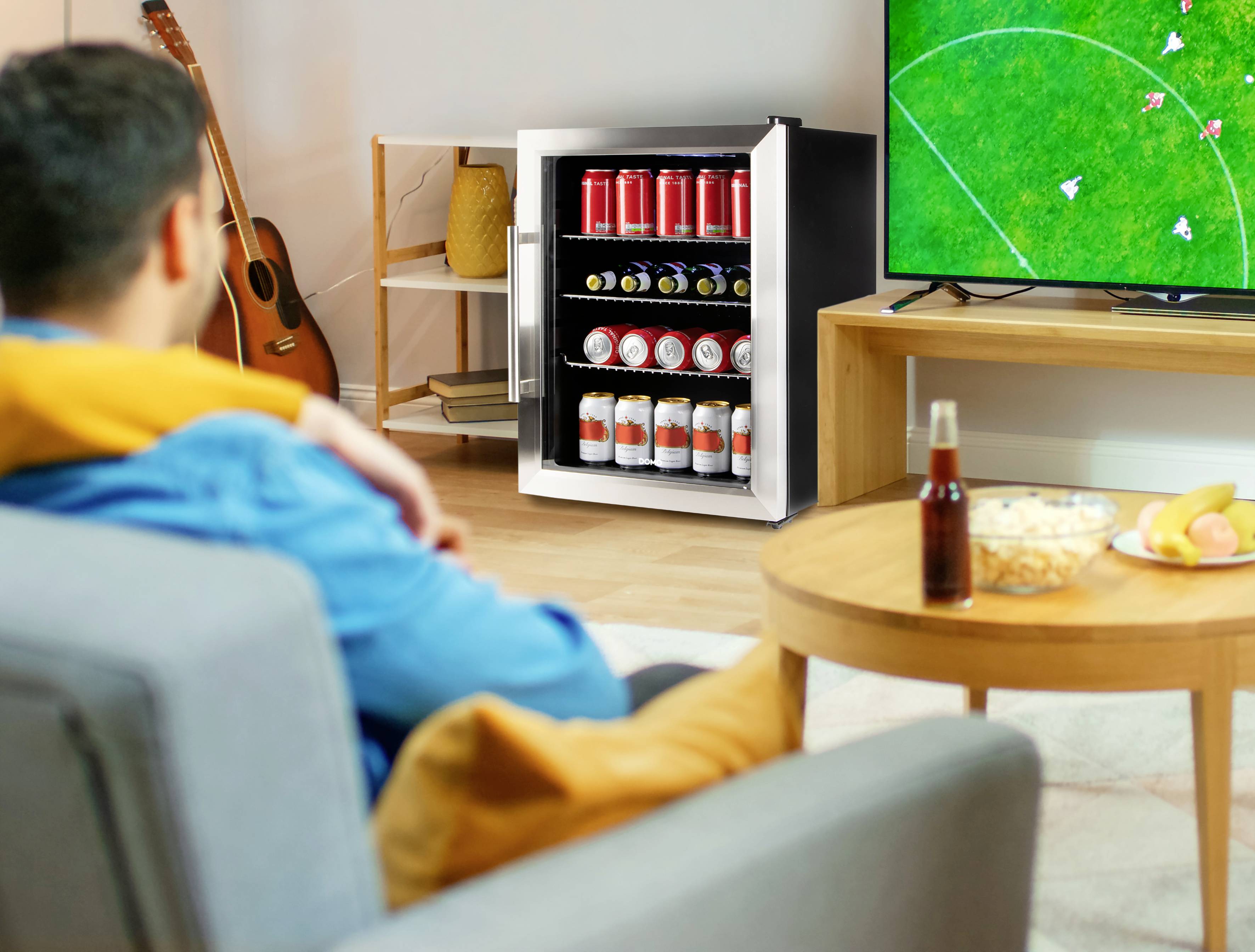 Ein Mann sitzt auf dem Sofa und schaut ein Fußballspiel im Fernsehen. Vor ihm steht ein Mini-Kühlschrank voller Getränke, daneben Snacks.