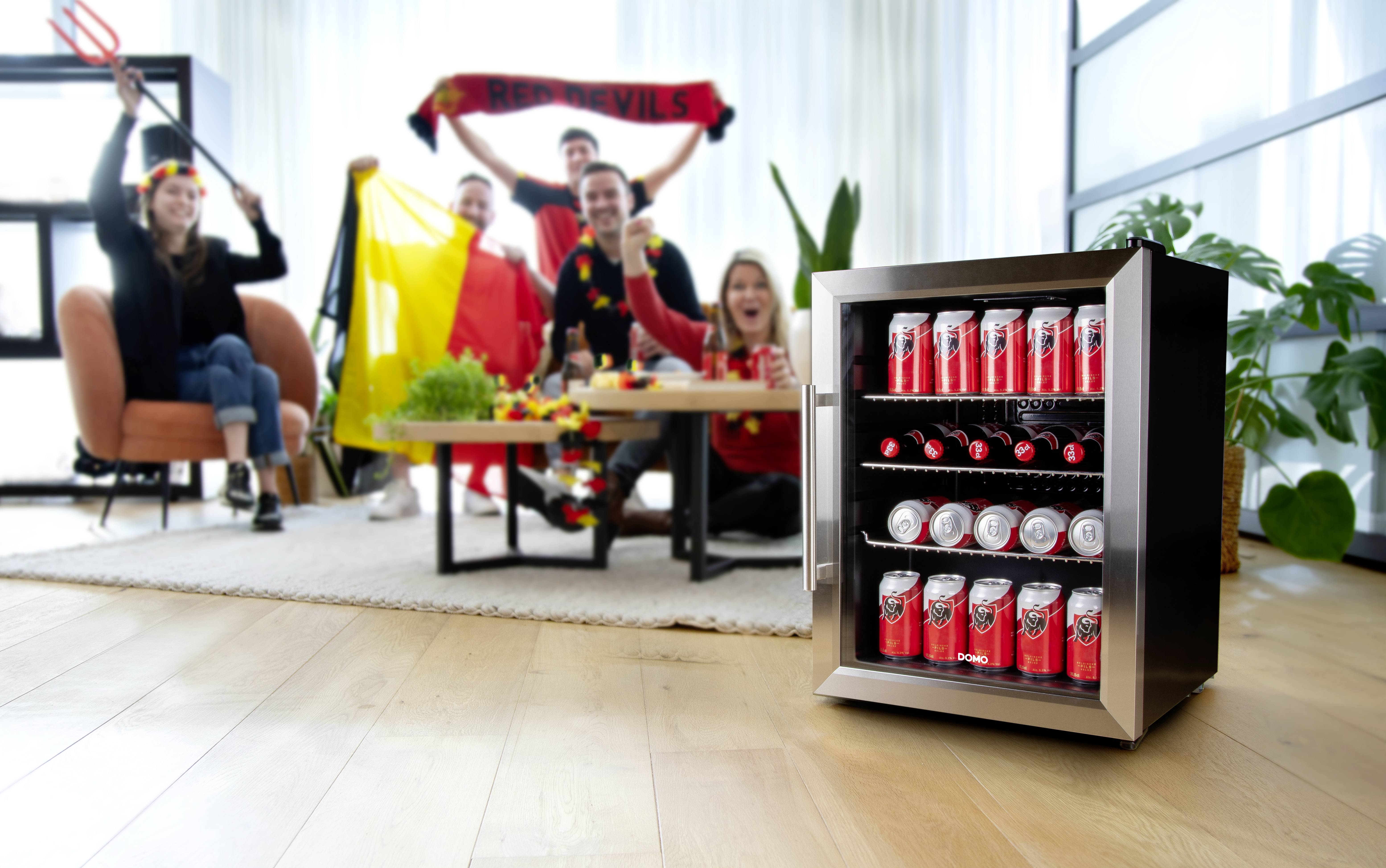 Eine Gruppe jubelt vor einem Tisch mit einer belgischen Flagge, während im Vordergrund ein kleiner Kühlschrank mit Getränkedosen steht.