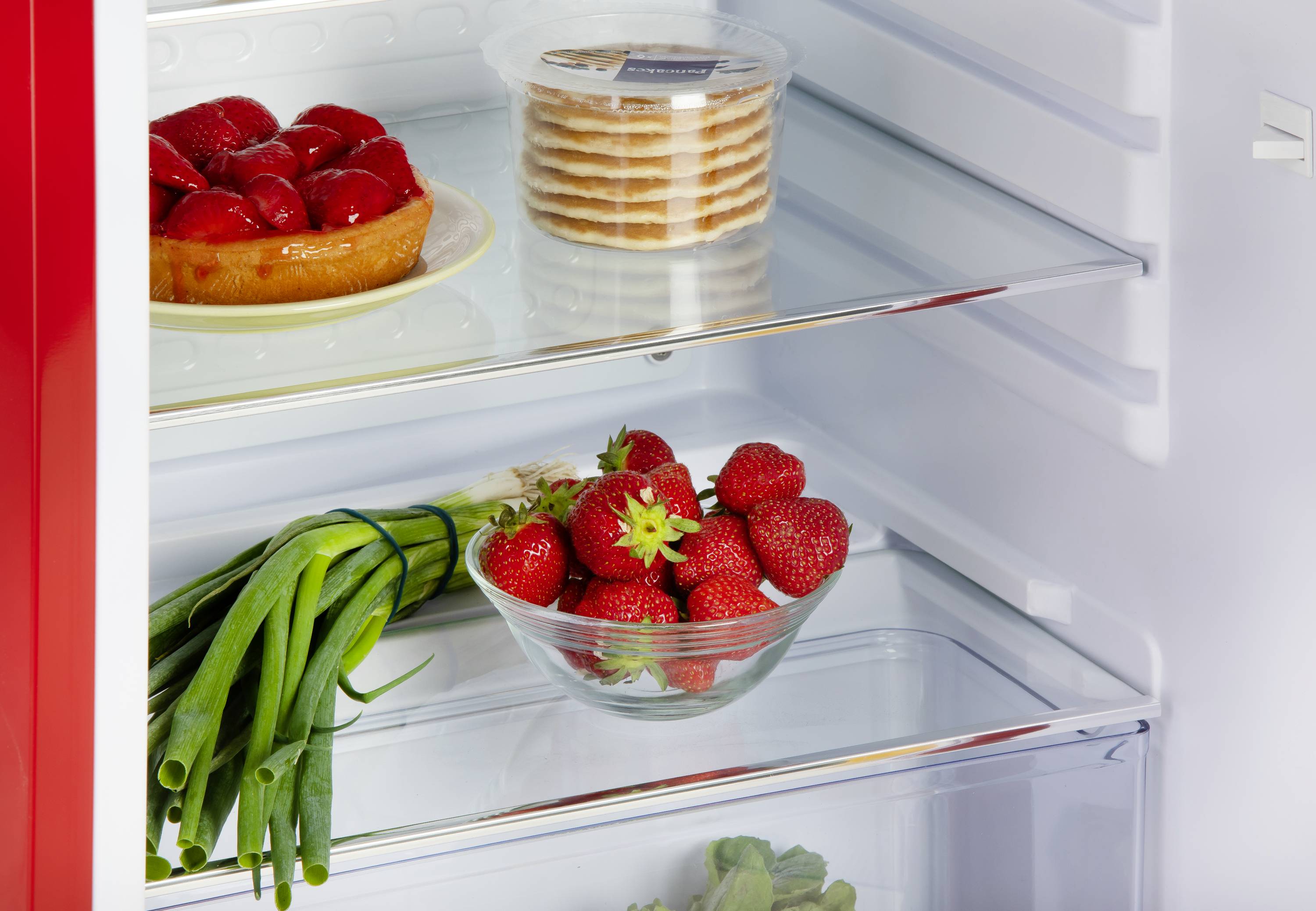 Ein offener Kühlschrank zeigt Torte, Kekse, Frühlingszwiebeln und Erdbeeren. Die Lebensmittel sind ordentlich auf Regalen angeordnet.