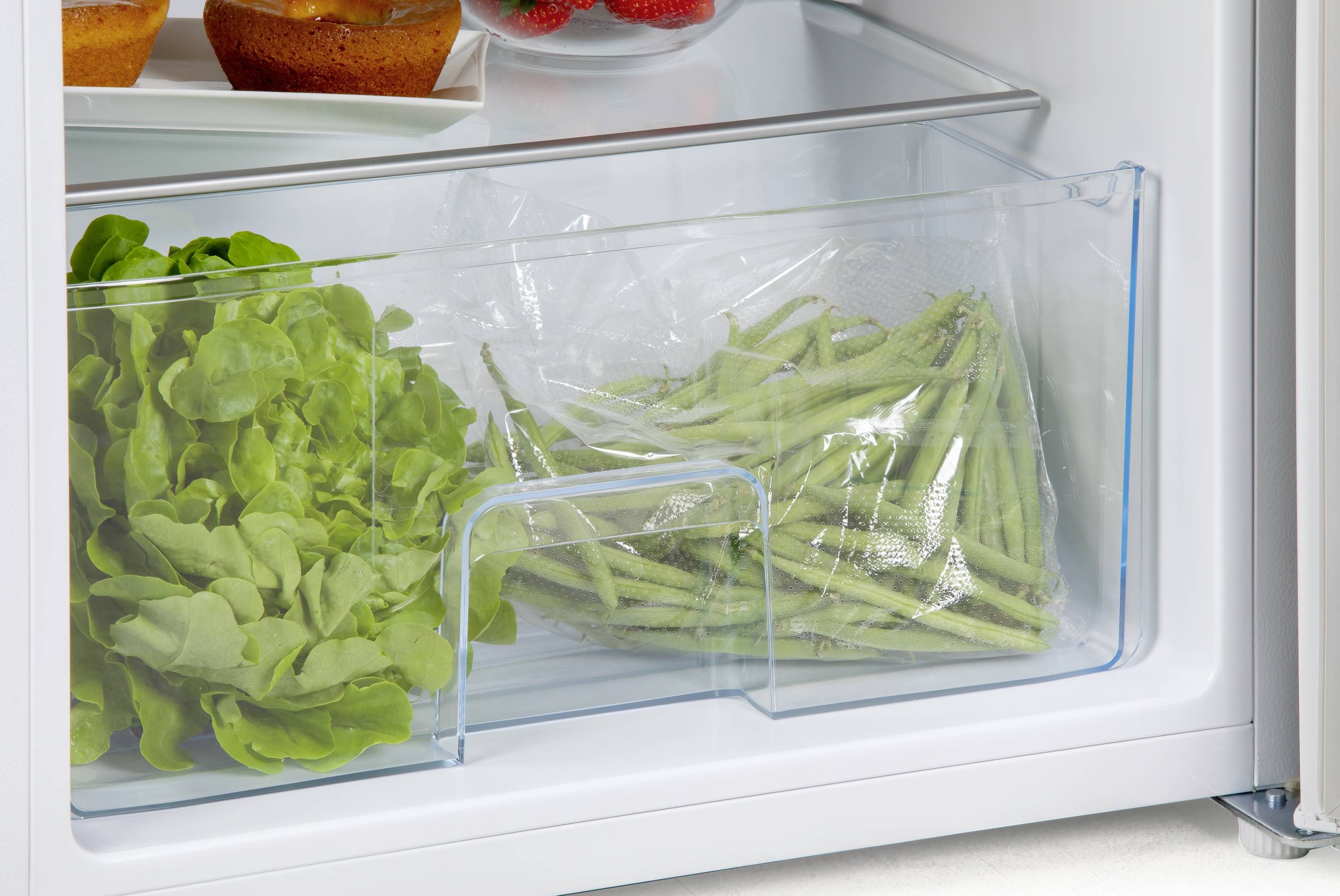 Ein Kühlschrank mit einem Gemüsefach, das frischen Salat und grüne Bohnen enthält. Im Hintergrund sind Muffins und Erdbeeren zu sehen.