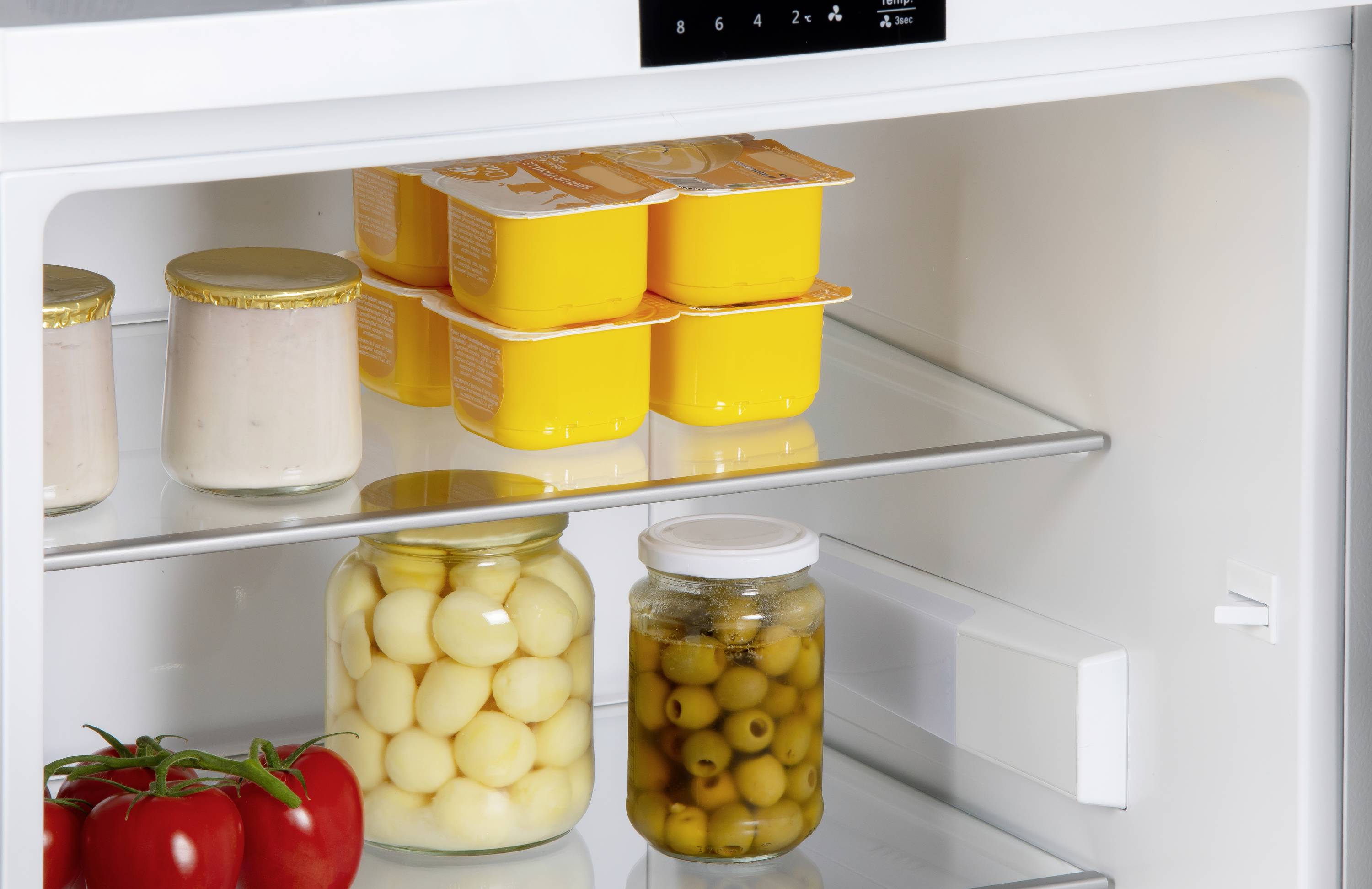 Im Kühlschrank befinden sich Tomaten, ein Glas mit weißen Eiern, ein Glas mit Oliven und acht gelbe Joghurtbecher.