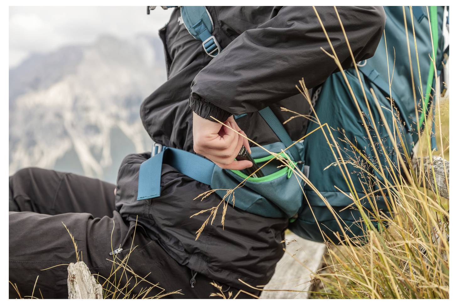 Person in a schwarzen Jacke verstellt den Hüftgurt eines türkisfarbenen Rucksacks, während er auf trockenen Gras mit Bergen im Hintergrund sitzt.