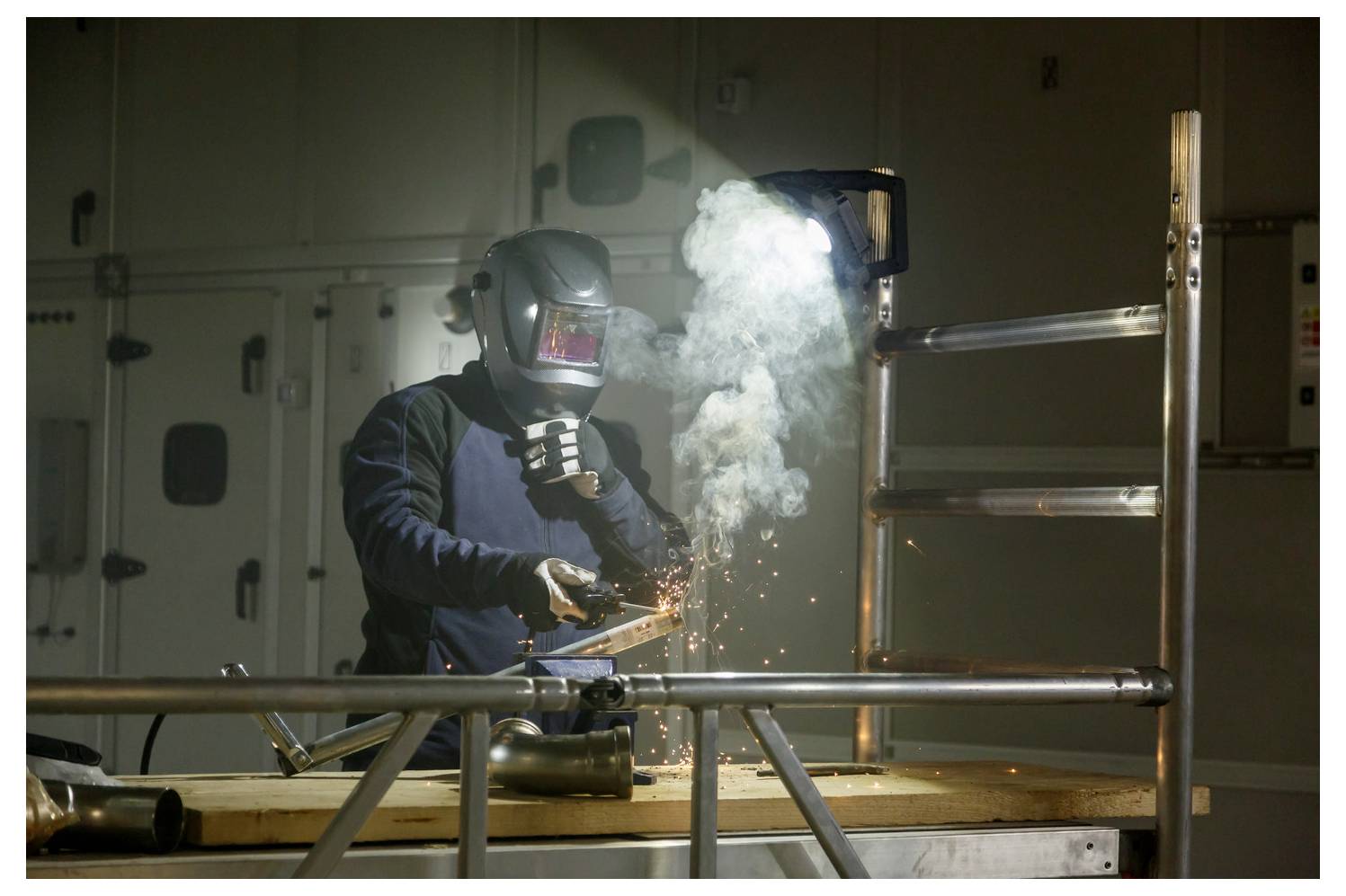 Ein Arbeiter mit Schweißhelm schweißt Metall in einer Industriewerkstatt, wobei helle Funken und Rauch um das Werkstück aufsteigen.