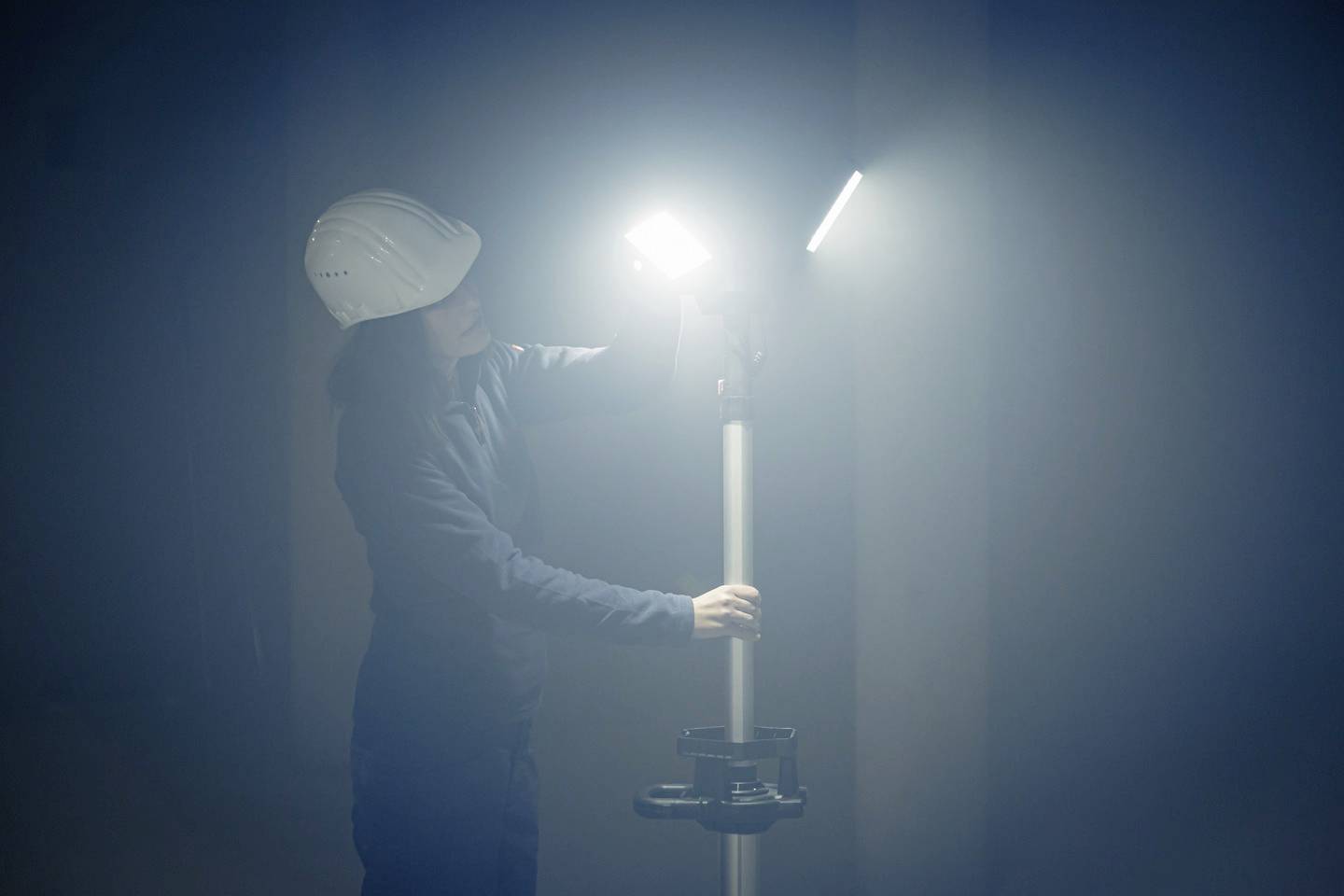 Eine Person mit Schutzhelm justiert ein helles, leuchtendes Baustellenlicht in einem nebligen Raum.