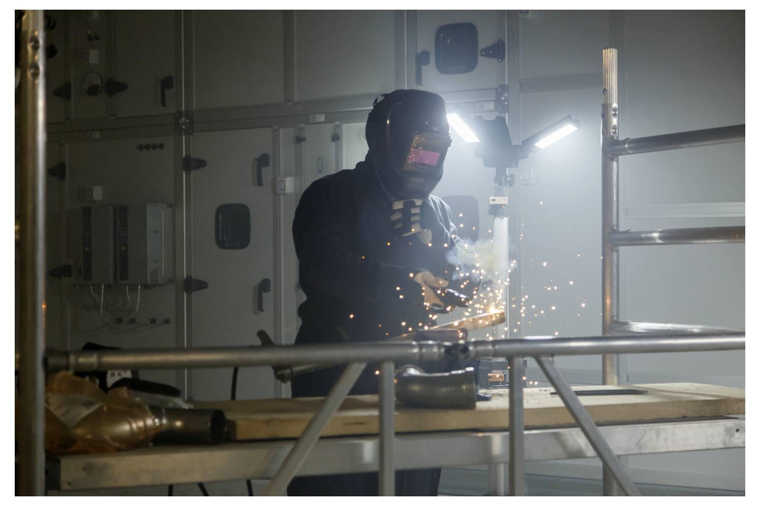 Arbeiter in Schutzausrüstung schweißt Metall in einem Innenraum, mit hellen Funken und Industrieausrüstung im Hintergrund.