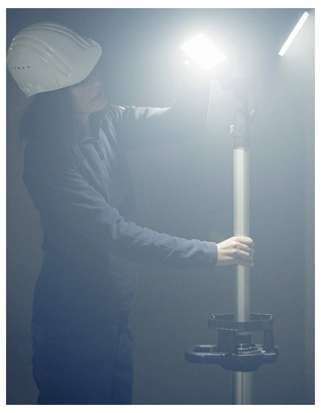 Arbeiter in einem Schutzhelm stellt ein helles Arbeitslicht auf einem Ständer in einer dunstig-industriellen Umgebung ein.