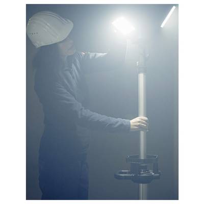 Arbeiter in einem Schutzhelm stellt ein helles Arbeitslicht auf einem Ständer in einer dunstig-industriellen Umgebung ein.