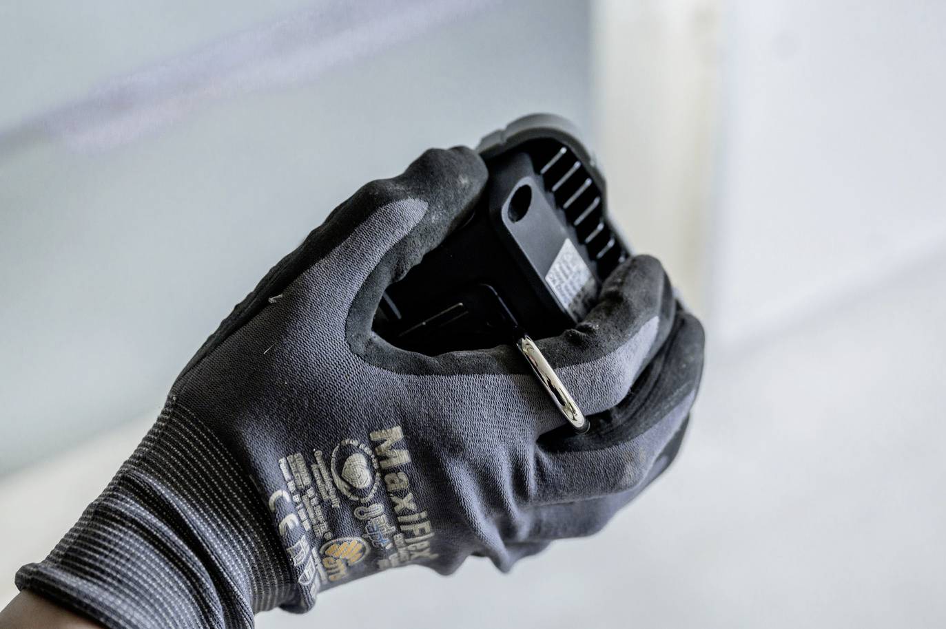 Eine Person hält mit einer schwarzen, gepolsterten Arbeitshandschuh einen schwarzen, rechteckigen Gegenstand mit einer kleinen Metallklammer.