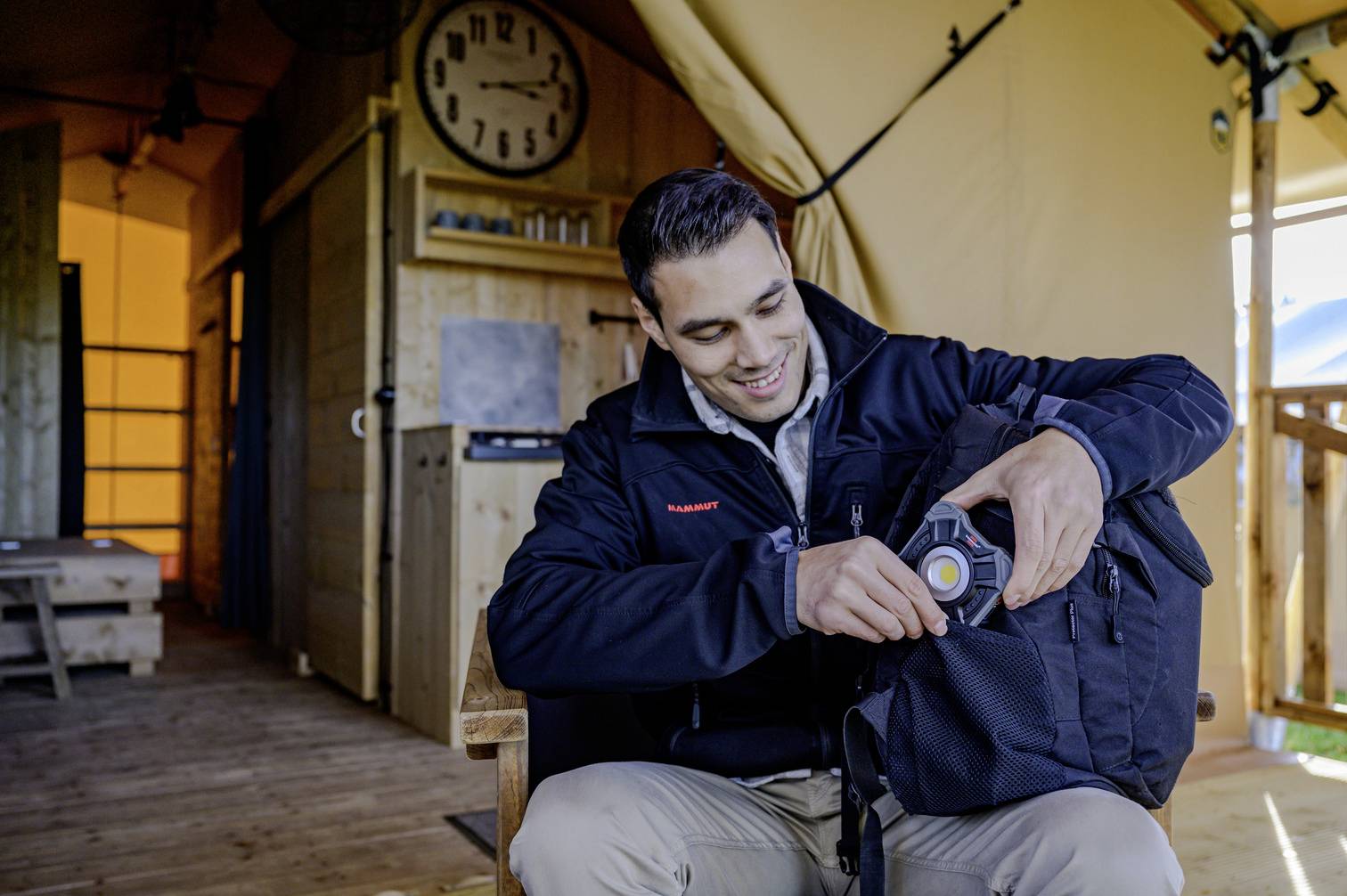 Ein Mann in einer Outdoor-Umgebung sitzt auf einem Stuhl und überprüft den Inhalt seines Rucksacks. Im Hintergrund befindet sich eine Holzhütte mit einer Uhr an der Wand.