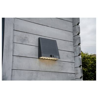 Schwarzes wandmontiertes Solarleuchte mit Bewegungsmelder und leuchtenden LEDs an einer grauen Holzaußenwand.