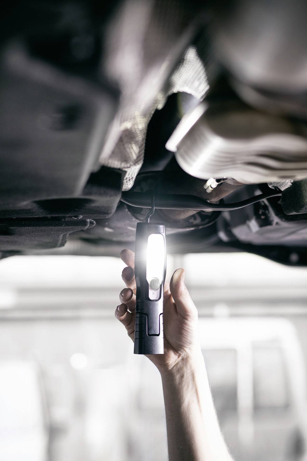 Eine Hand hält eine leuchtende Taschenlampe unter einem Auto, vermutlich für eine Inspektion oder Reparatur.