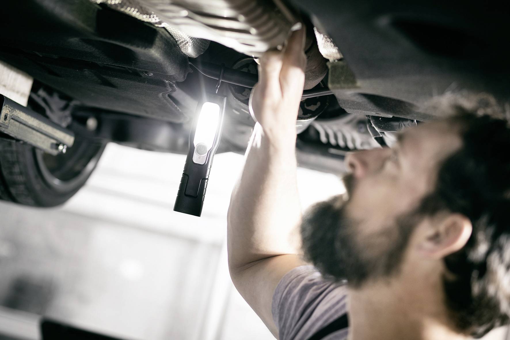 Ein Mechaniker überprüft unter einem Fahrzeug einen Bauteil. Eine Lampe leuchtet den Arbeitsbereich aus.