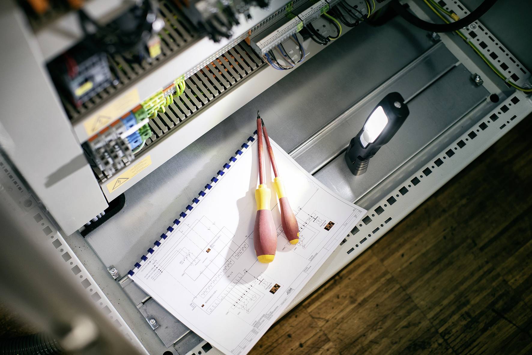 Ein geöffneter Schaltschrank enthält Stromkreispläne, zwei Schraubenzieher und eine Taschenlampe, die gerichtete Arbeiten suggerieren.
