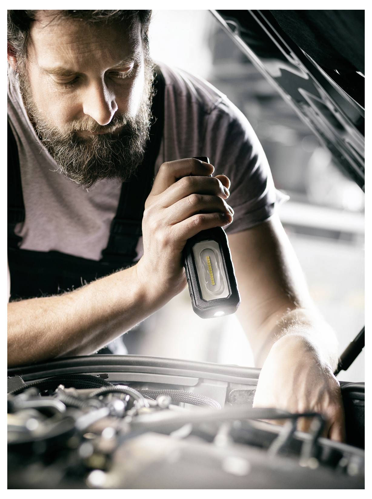 Mechaniker, der einen Automotor unter der offenen Motorhaube mit einer tragbaren Arbeitsleuchte inspiziert.