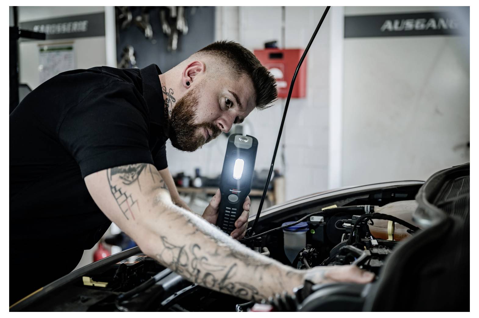 Mechaniker überprüft einen Automotor in einer Werkstatt mit einer tragbaren Taschenlampe, lehnt sich über die offene Motorhaube.