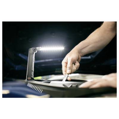 Die Hände eines Mechanikers benutzen ein Werkzeug unter einem hellen Arbeitslicht in einem Motorraum während einer Fahzeugreparatur.