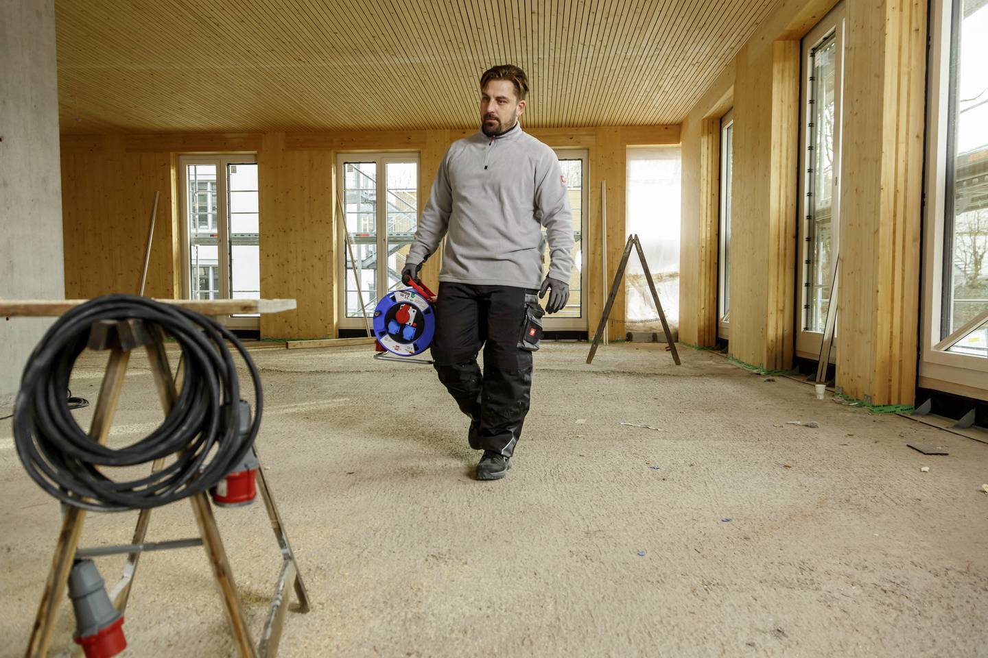 Ein Bauarbeiter trägt eine Kabeltrommel durch einen hellen Raum mit Holzverkleidung und großen Fenstern. Im Hintergrund stehen Leitern.