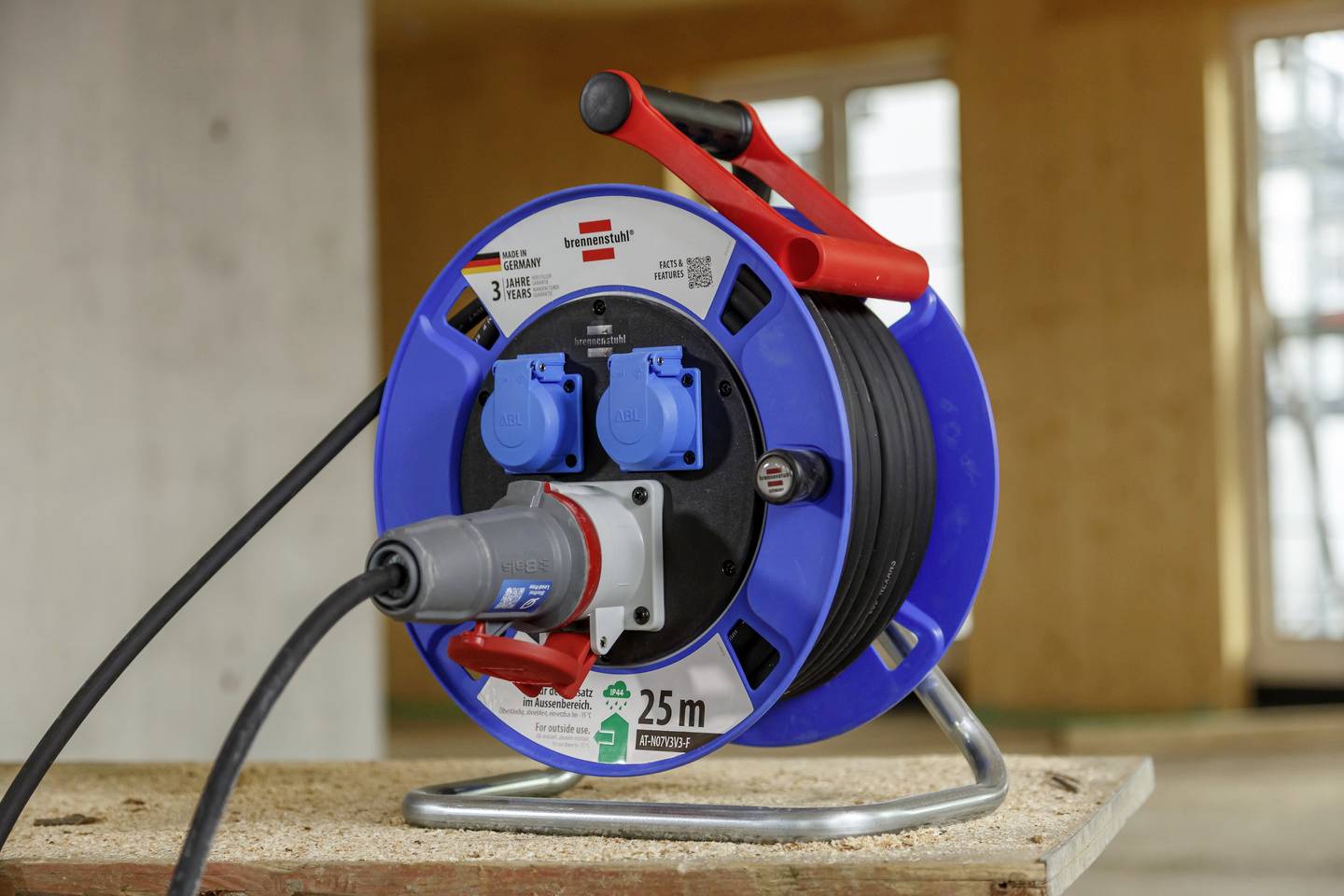 Ein blaues Verlängerungskabel auf einer Baustelle, aufgerollt auf einer Trommel mit drei blauen Steckdosen und einem roten Griff.