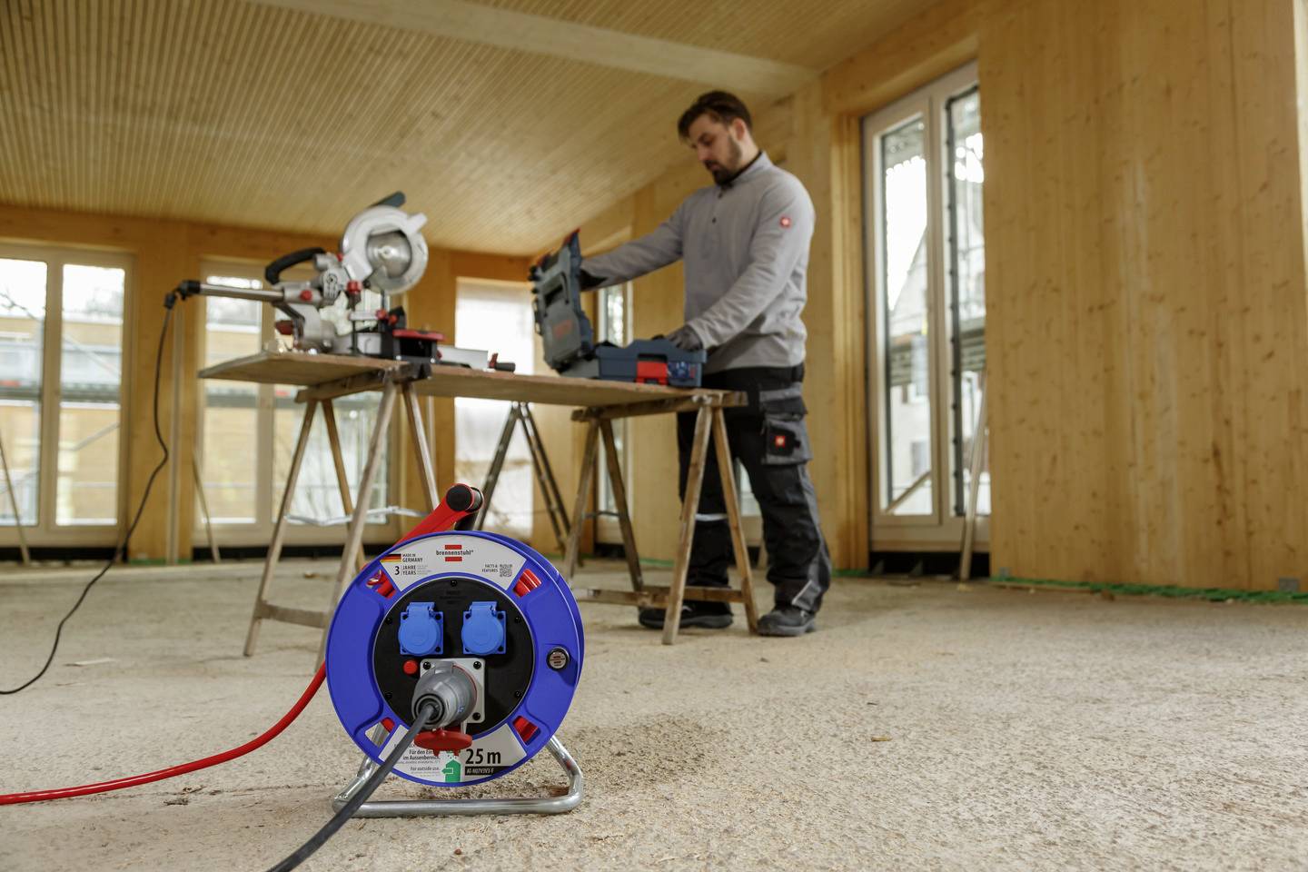 Ein Handwerker steht in einem unfertigen Raum und bedient eine Säge auf einem Tisch. Im Vordergrund ist eine Kabeltrommel zu sehen.