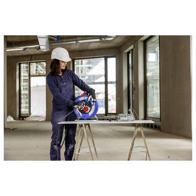 Bauarbeiter in blauem Schutzanzug und weißem Schutzhelm, der eine Elektrosäge auf einem Brett in einem unvollendeten Gebäudeinneren benutzt.
